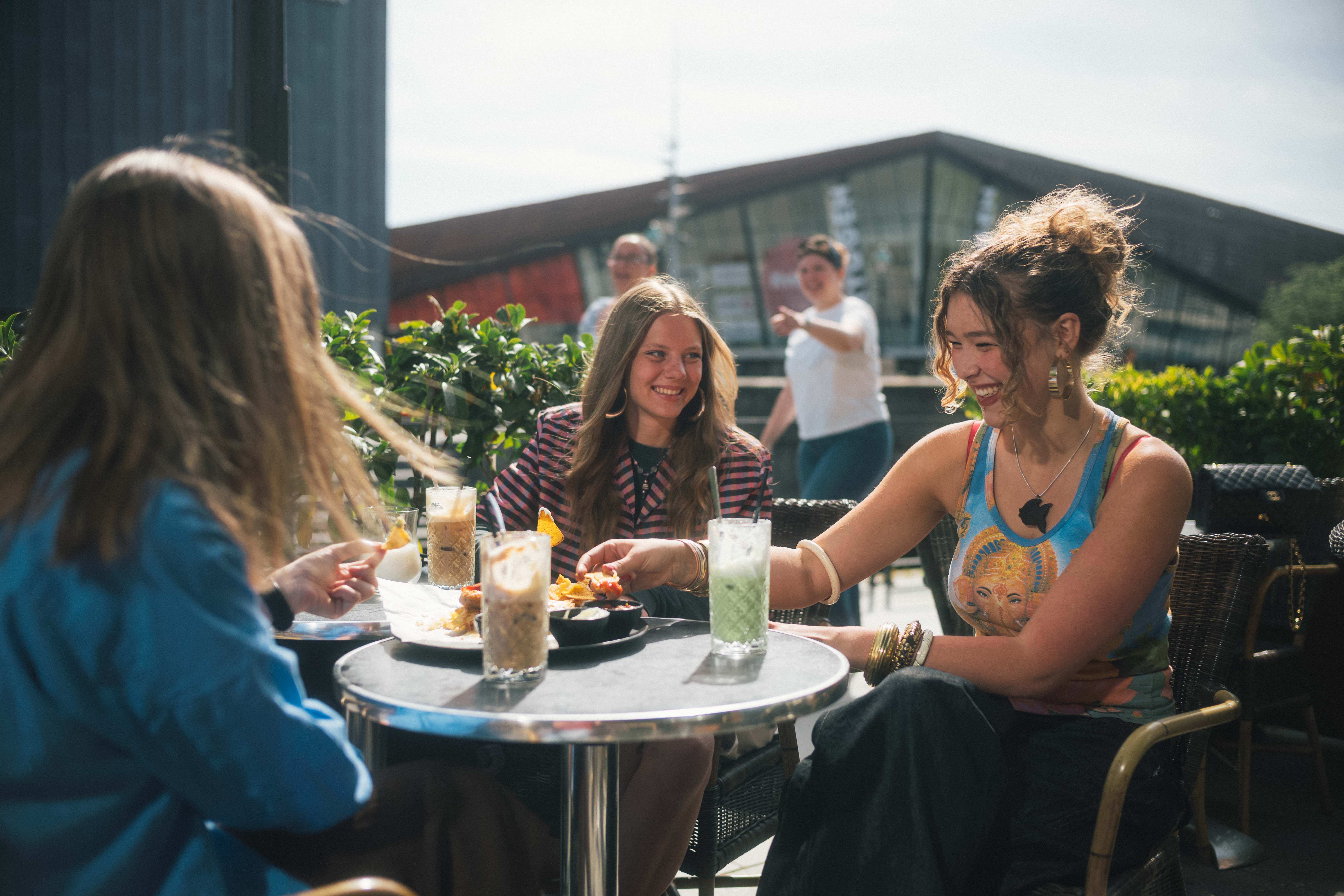 Tre unge kvinder sidder udenfor en café og snakker. Der er med og drikke på bordene imellem dem.