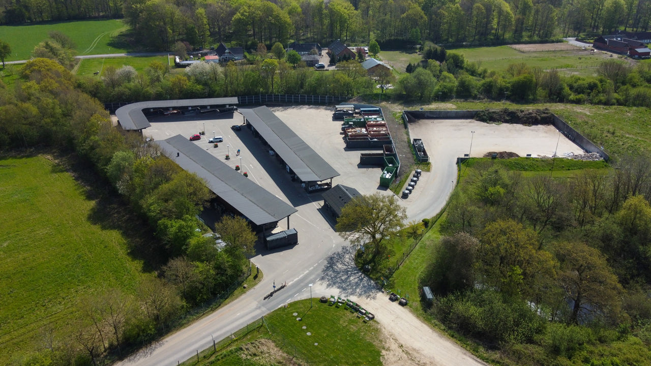Luftfoto af Børkop Genbrugsplads omgivet af grønne træer og landlige omgivelser. Pladsen er inddelt i overdækkede sorteringsområder og containere til forskellige affaldstyper.