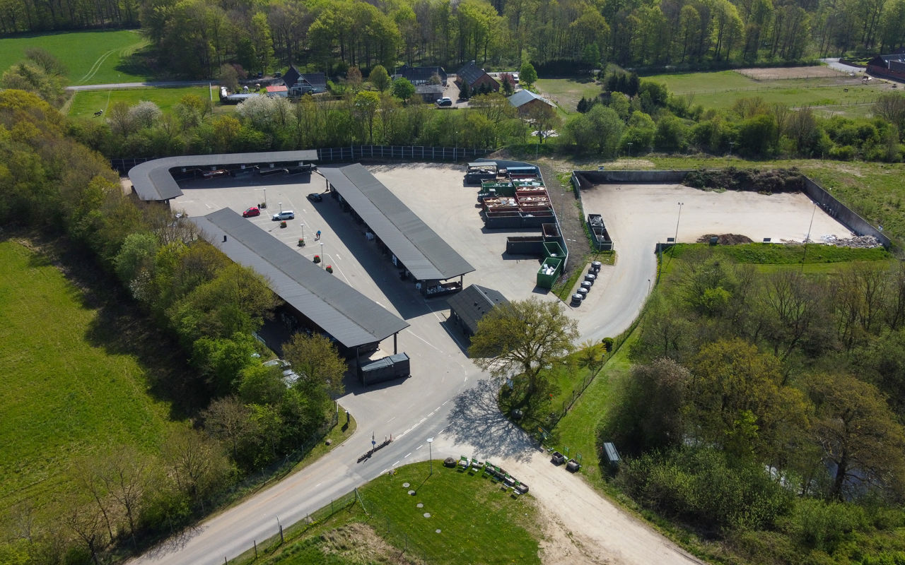 Luftfoto af Børkop Genbrugsplads omgivet af grønne træer og landlige omgivelser. Pladsen er inddelt i overdækkede sorteringsområder og containere til forskellige affaldstyper.