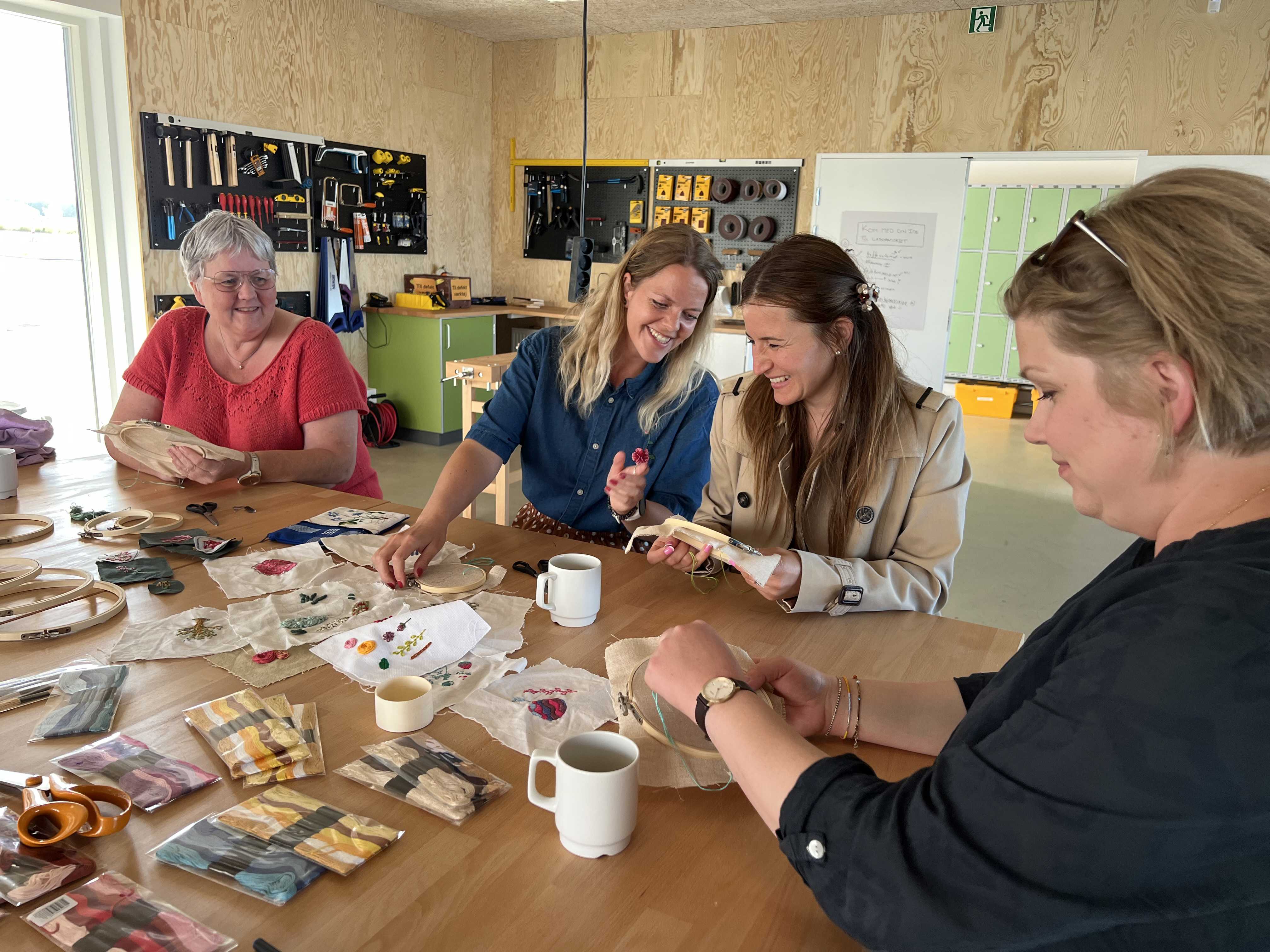 Fire smilede kvinder i alle aldre sidder ved et arbejdsbord i Laboratoriet på RCV og broderer.