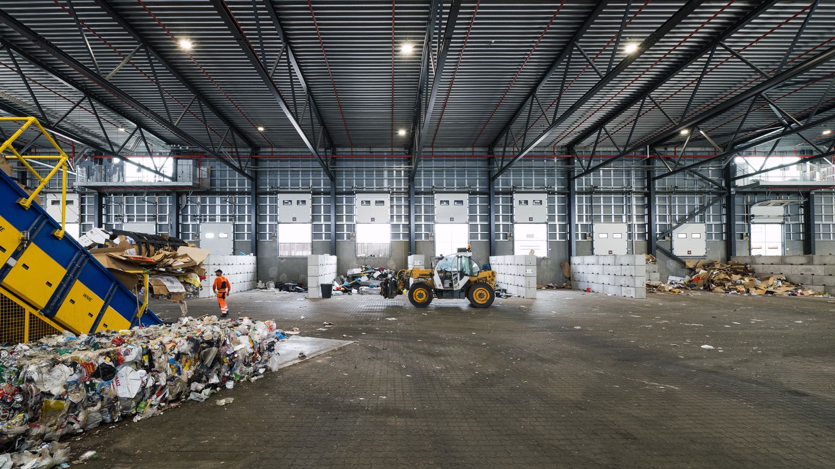 Indendørs foto af RessourceCenter Vejle's affaldssorteringshal med en gaffeltruck i arbejde og store bunker af sorteret affald, klar til videre forarbejdnin