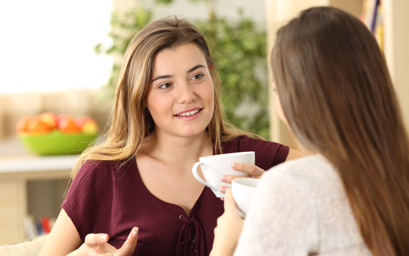 To piger i en samtale siddende tæt på hinanden med kaffekopper i hænderne