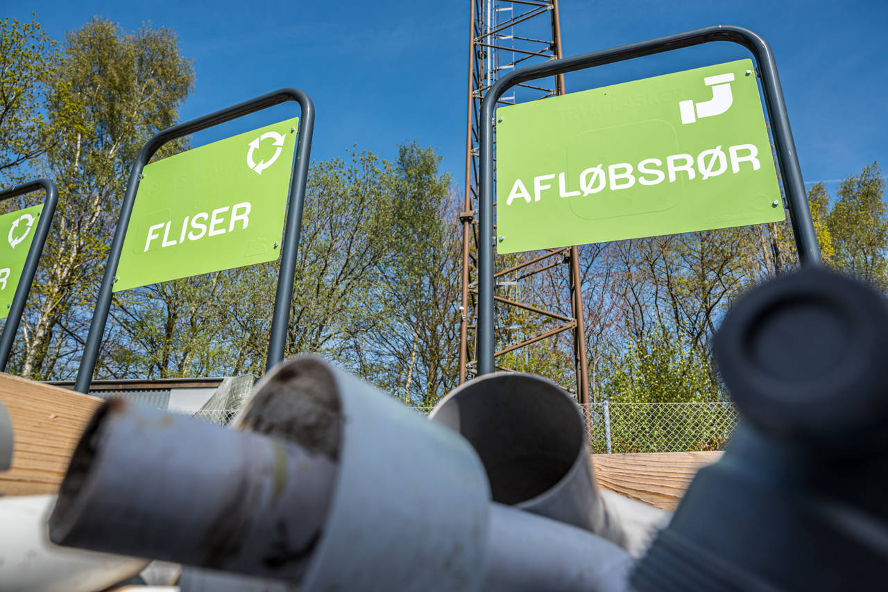 Grønt skilt med teksten 'FLISER' og 'AFLØBSRØR' over en palle med overskudsmaterialer på BygIgen-området ved Egtved Genbrugsplads.