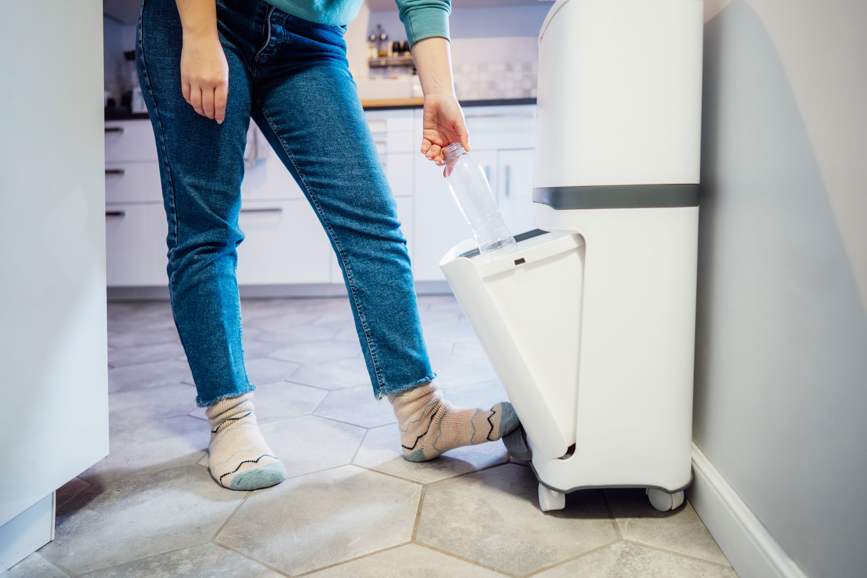 Person iført jeans og uldsokker sorterer en plastflaske i en hvid affaldsspand med flere rum i et moderne køkken. Personen åbner affaldsspanden med en fodpedal.
