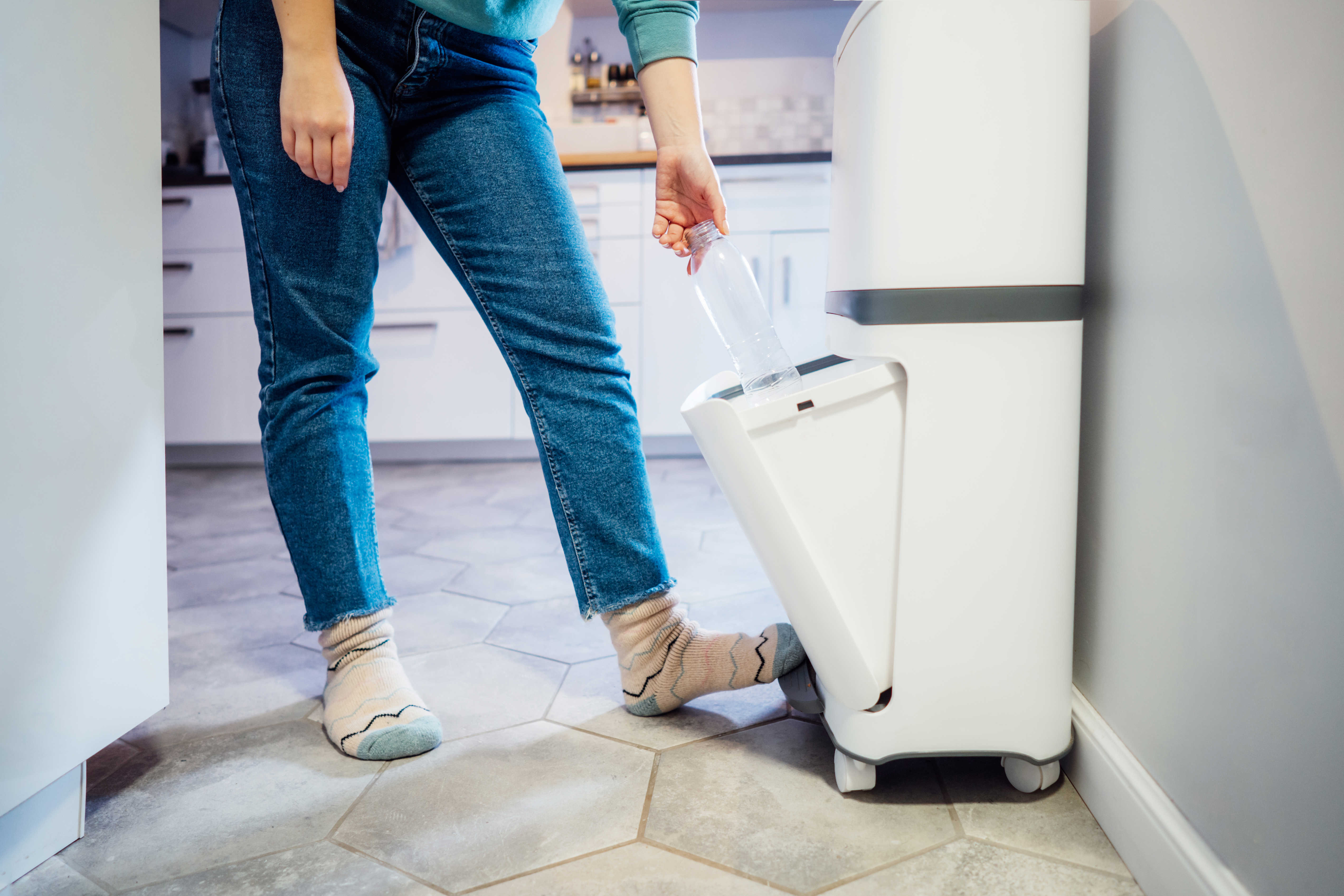 Person iført jeans og uldsokker sorterer en plastflaske i en hvid affaldsspand med flere rum i et moderne køkken. Personen åbner affaldsspanden med en fodpedal.