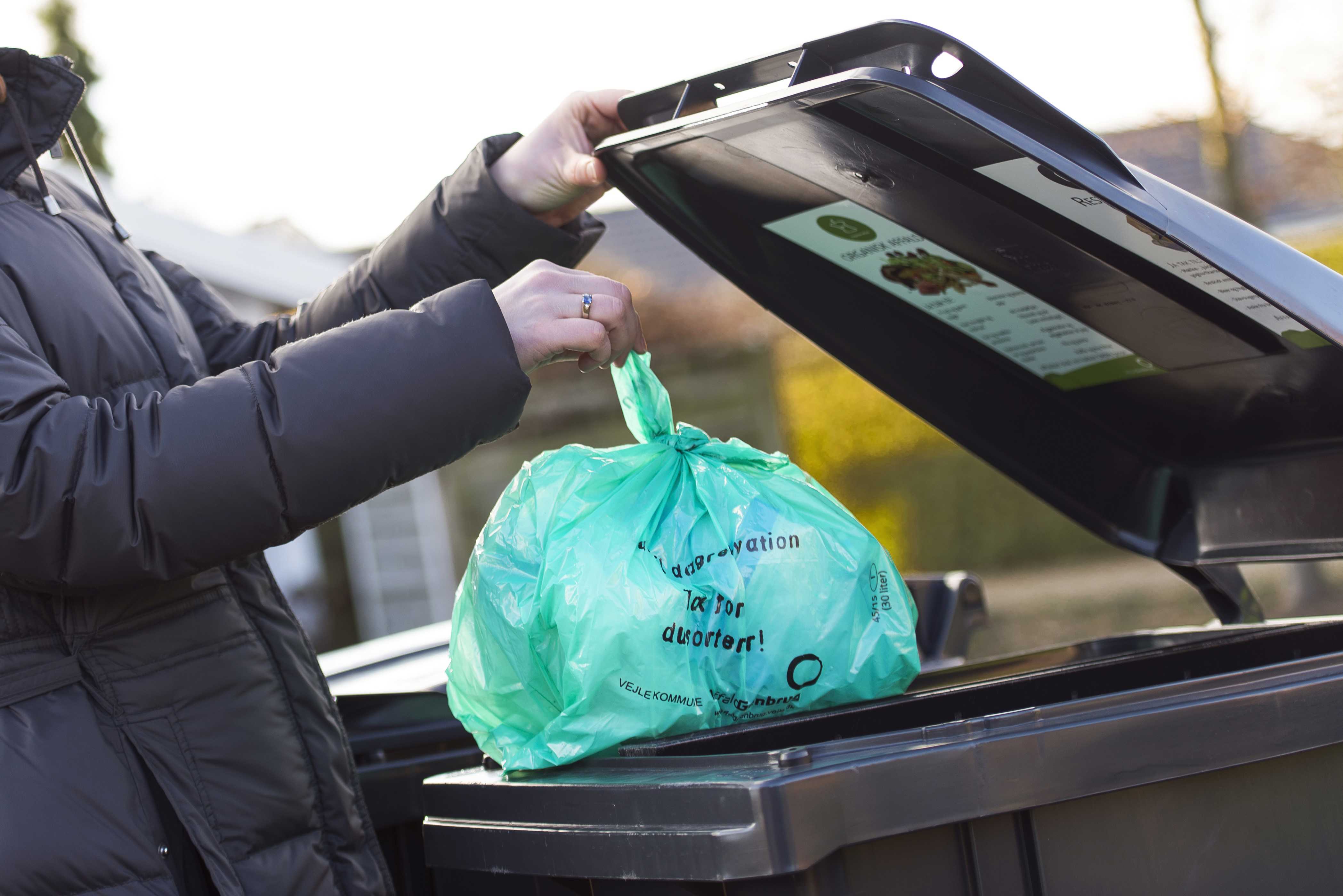 erson iført mørk jakke smider en grøn biologisk nedbrydelig pose med madaffald i en sort affaldscontainer. Containerlåget har en informationsplakat om korrekt affaldssortering. Baggrunden viser en villavej med hække og huse i sollys.