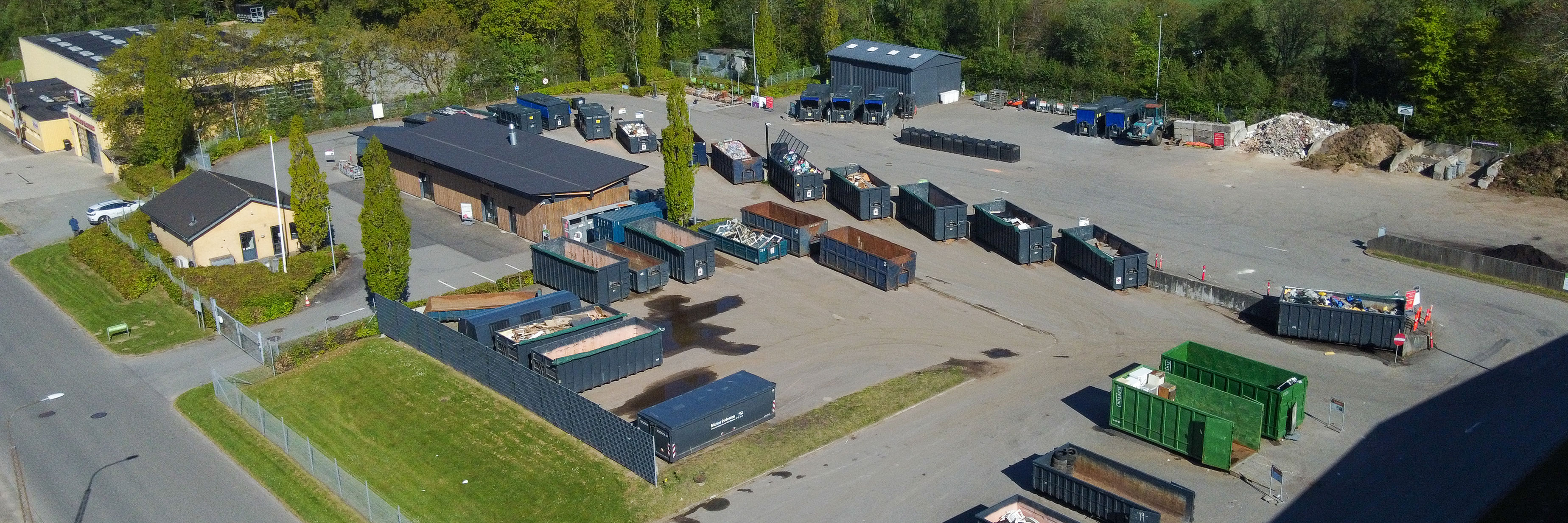 Oversigtsfoto af Egtved Genbrugsplads med udsyn til containere og sorteringsområder. Pladsen er omgivet af grønne træer og bygninger, og der er tydelige kørselsbaner til biler.