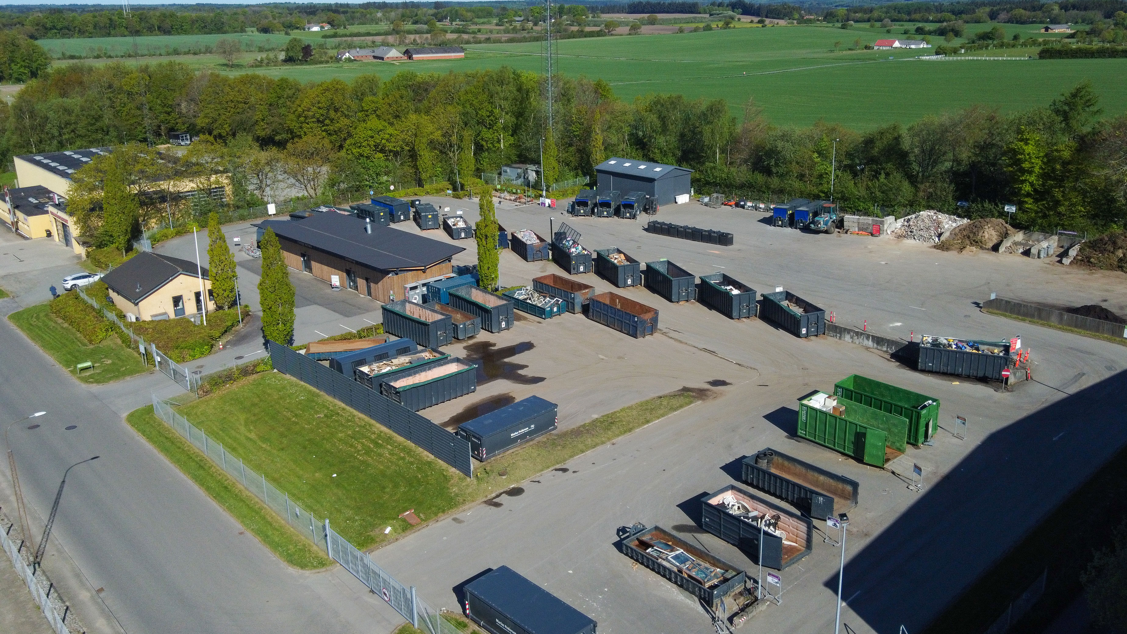 Oversigtsfoto af Egtved Genbrugsplads med udsyn til containere og sorteringsområder. Pladsen er omgivet af grønne træer og bygninger, og der er tydelige kørselsbaner til biler.