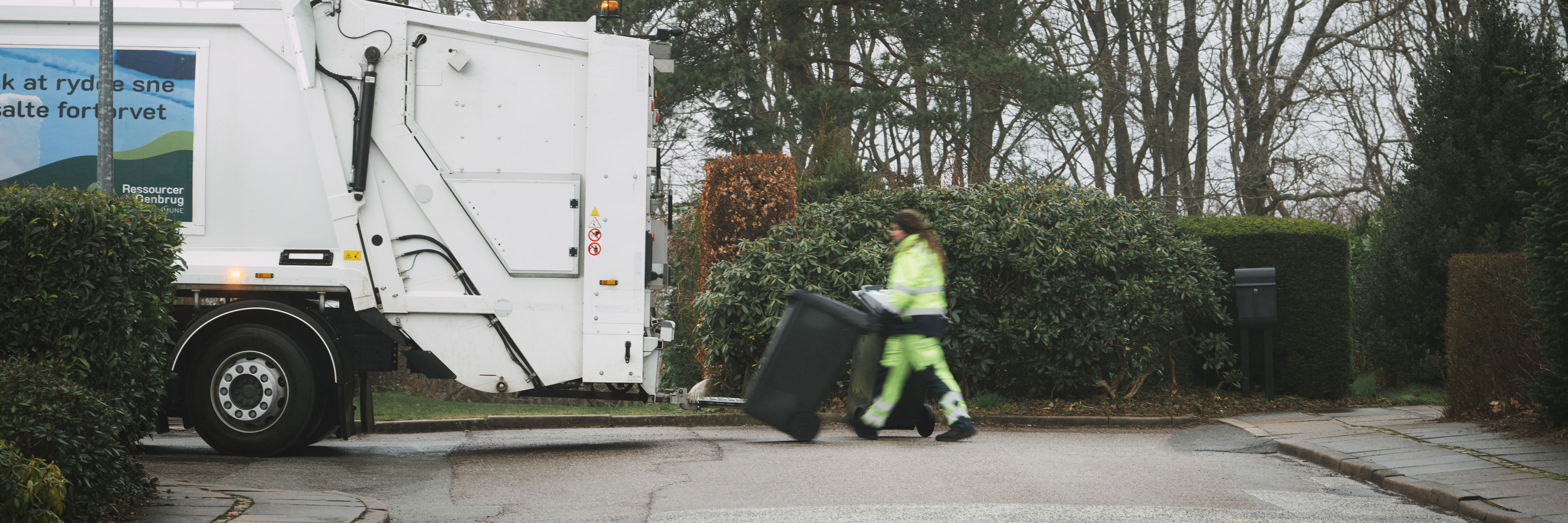 Skraldemand trækker en affaldsbeholder over vejen til skraldebilen på en stille villavej