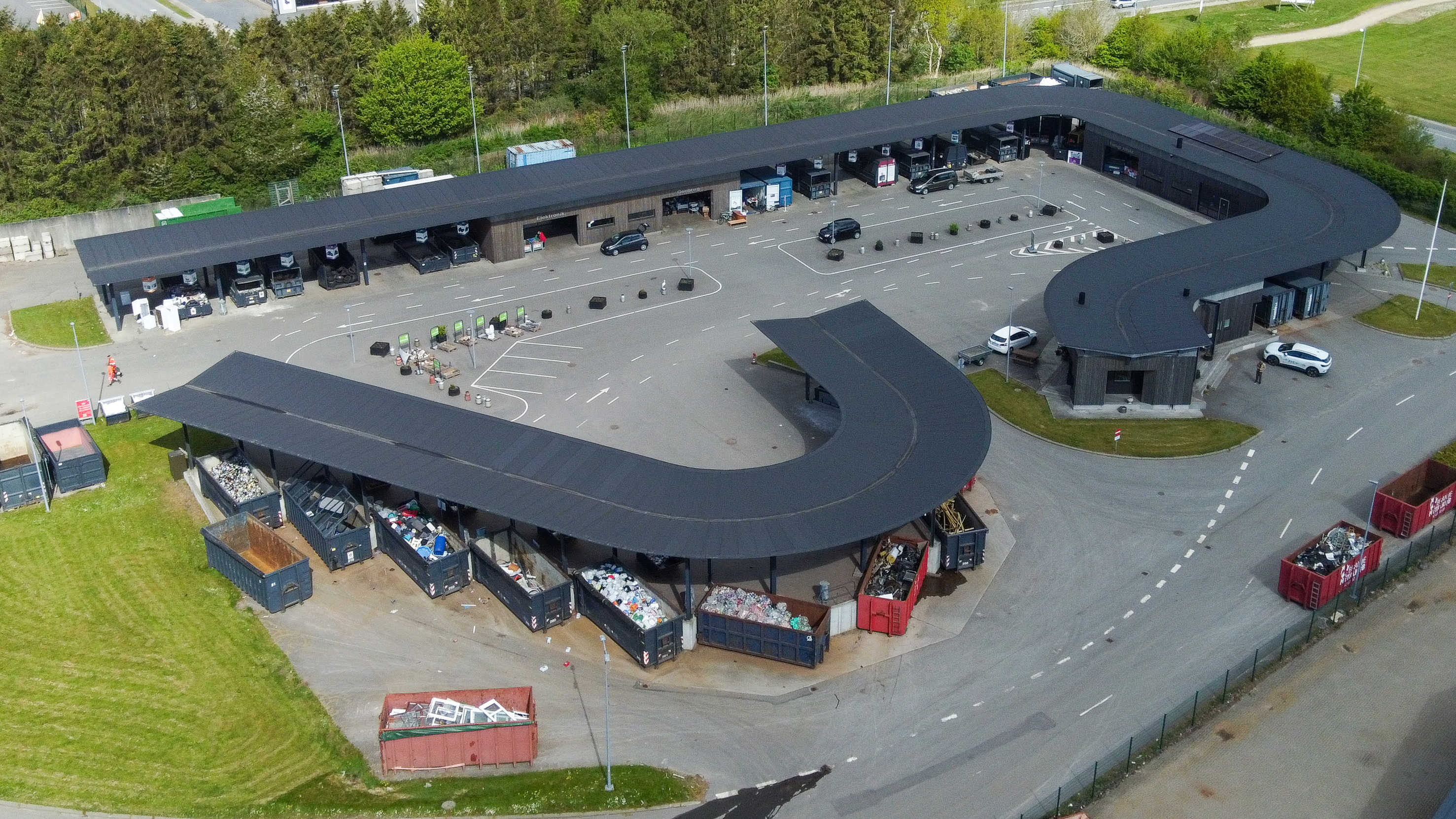 Oversigtsfoto af hele Give Genbrugsplads omgivet af grønne områder og vejnet. Billedet viser hele den buede bygning, kørselsretninger, containere og parkeringsområde.