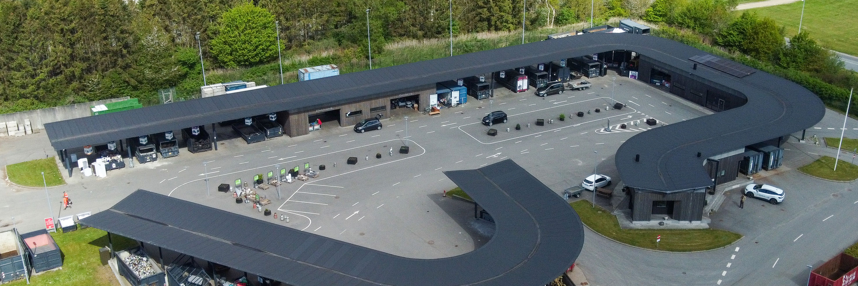 Oversigtsfoto af hele Give Genbrugsplads omgivet af grønne områder og vejnet. Billedet viser hele den buede bygning, kørselsretninger, containere og parkeringsområde.