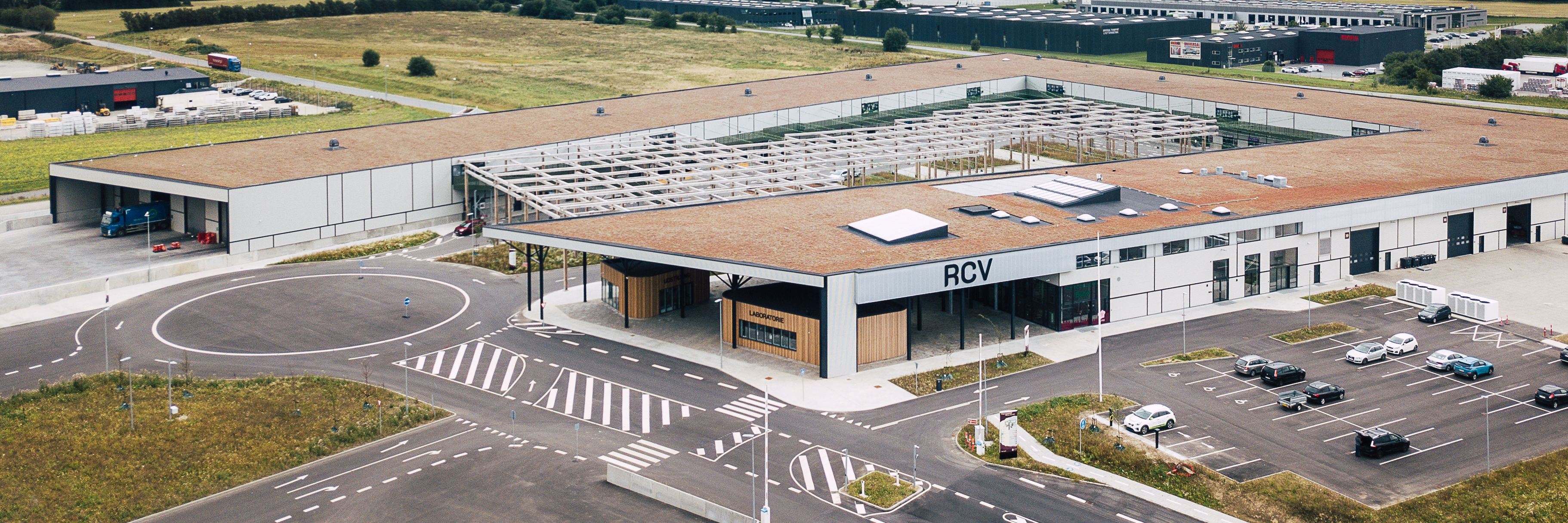 Luftfoto af RessourceCenter Vejle (RCV). Centret har en stor bygning med fladt tag, en parkeringsplads og tydeligt markerede vejbaner og indkørsler. Taget har solpaneler, og der er en åben konstruktion med trærammer i gårdområdet.