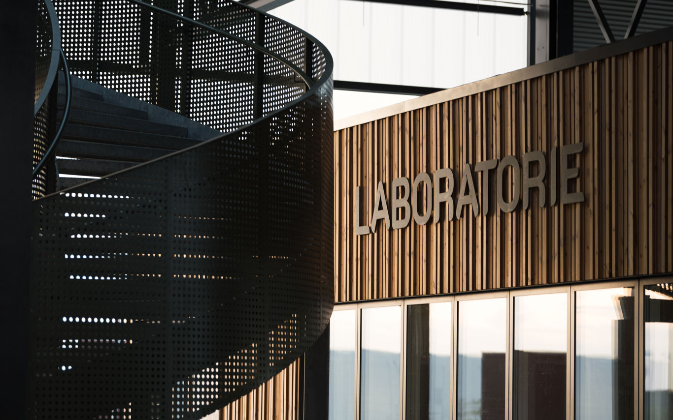 Nærbillede af facaden til Laboratoriet på RessourceCenter Vejle (RCV). Facaden er lavet af trælameller, og ordet "LABORATORIE" er monteret i store bogstaver. Til venstre ses en moderne vindeltrappe i perforeret metal, der tilføjer et industrielt præg til designet.