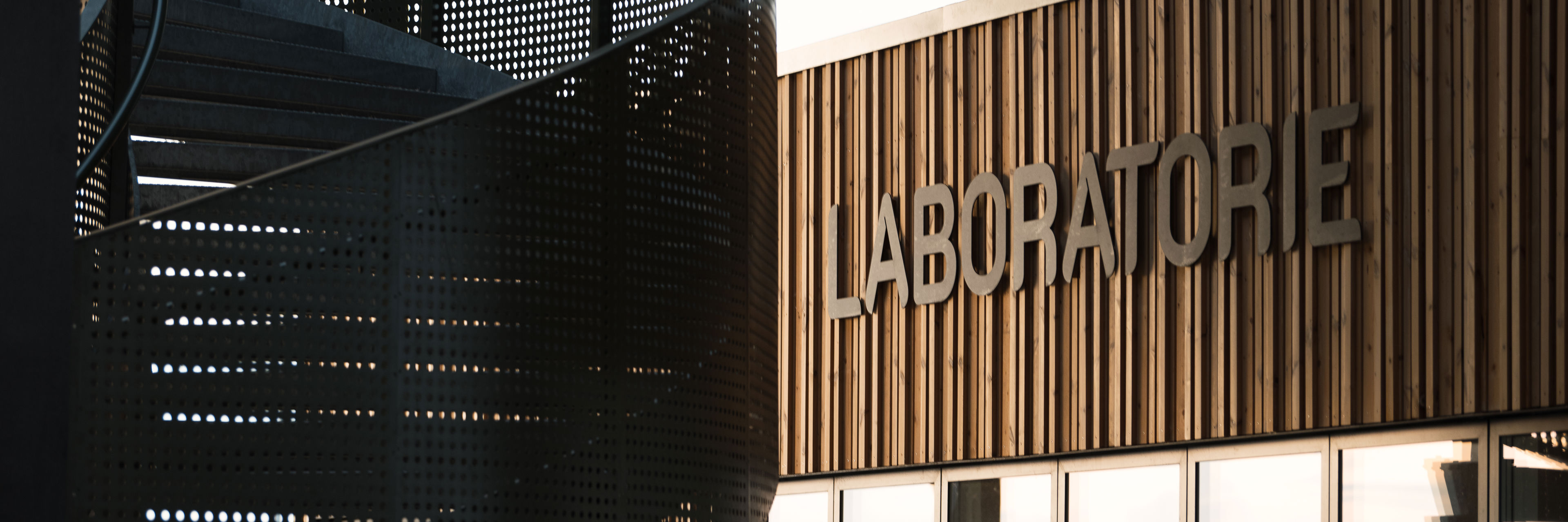 Nærbillede af facaden til Laboratoriet på RessourceCenter Vejle (RCV). Facaden er lavet af trælameller, og ordet "LABORATORIE" er monteret i store bogstaver. Til venstre ses en moderne vindeltrappe i perforeret metal, der tilføjer et industrielt præg til designet.