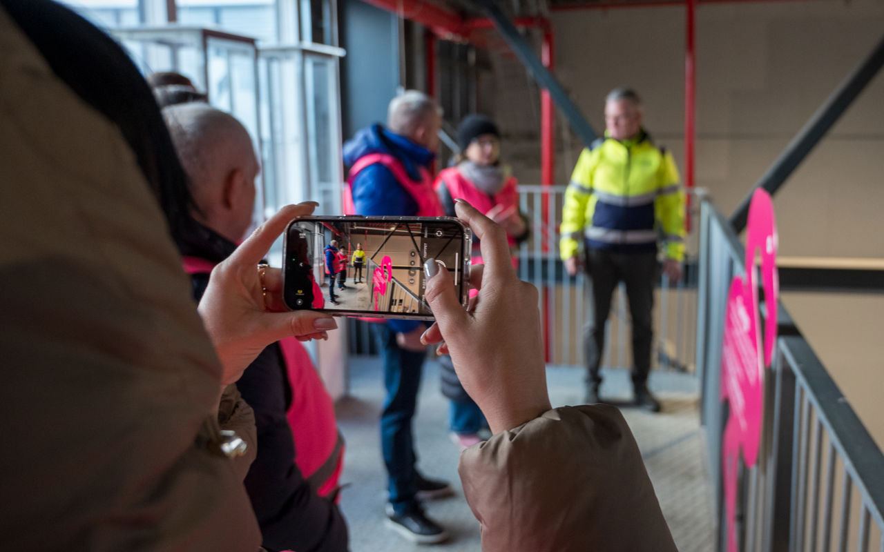 En person er i gang med at fotografere med en smartphone. På skærmen og i baggrunden ses en af platformene på Læringsbroen og flere personer i ført pink sikkerhedsveste står og taler sammen med en person i gul sikkerhedsjakke.