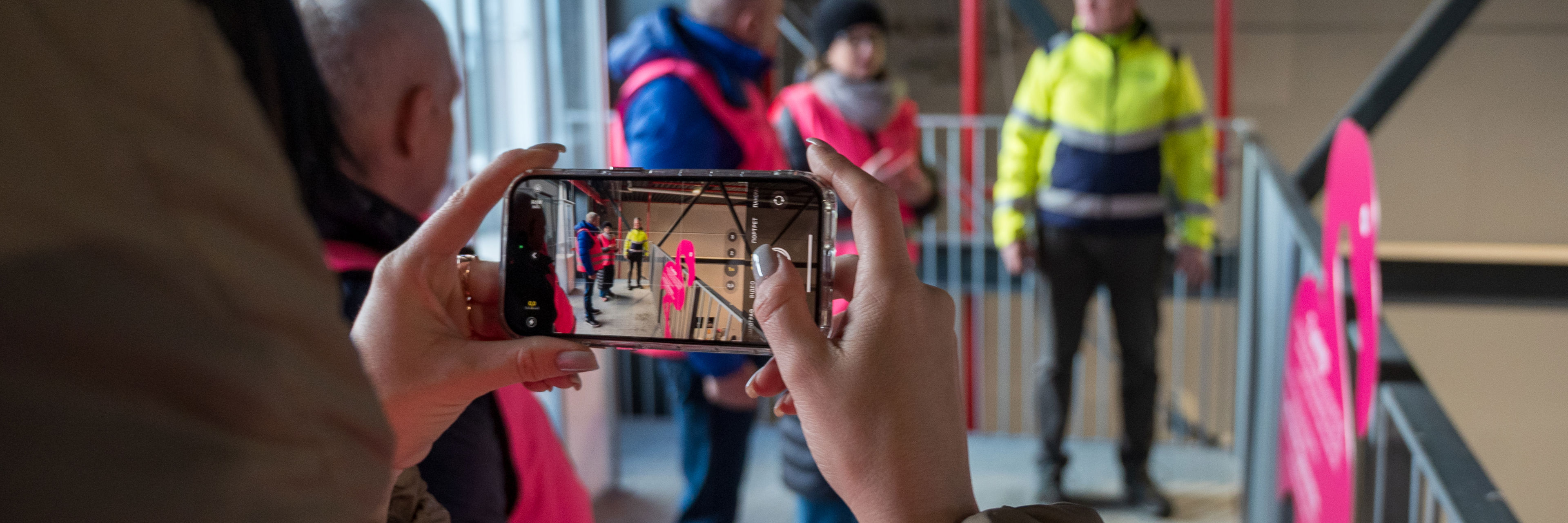En person er i gang med at fotografere med en smartphone. På skærmen og i baggrunden ses en af platformene på Læringsbroen og flere personer i ført pink sikkerhedsveste står og taler sammen med en person i gul sikkerhedsjakke.