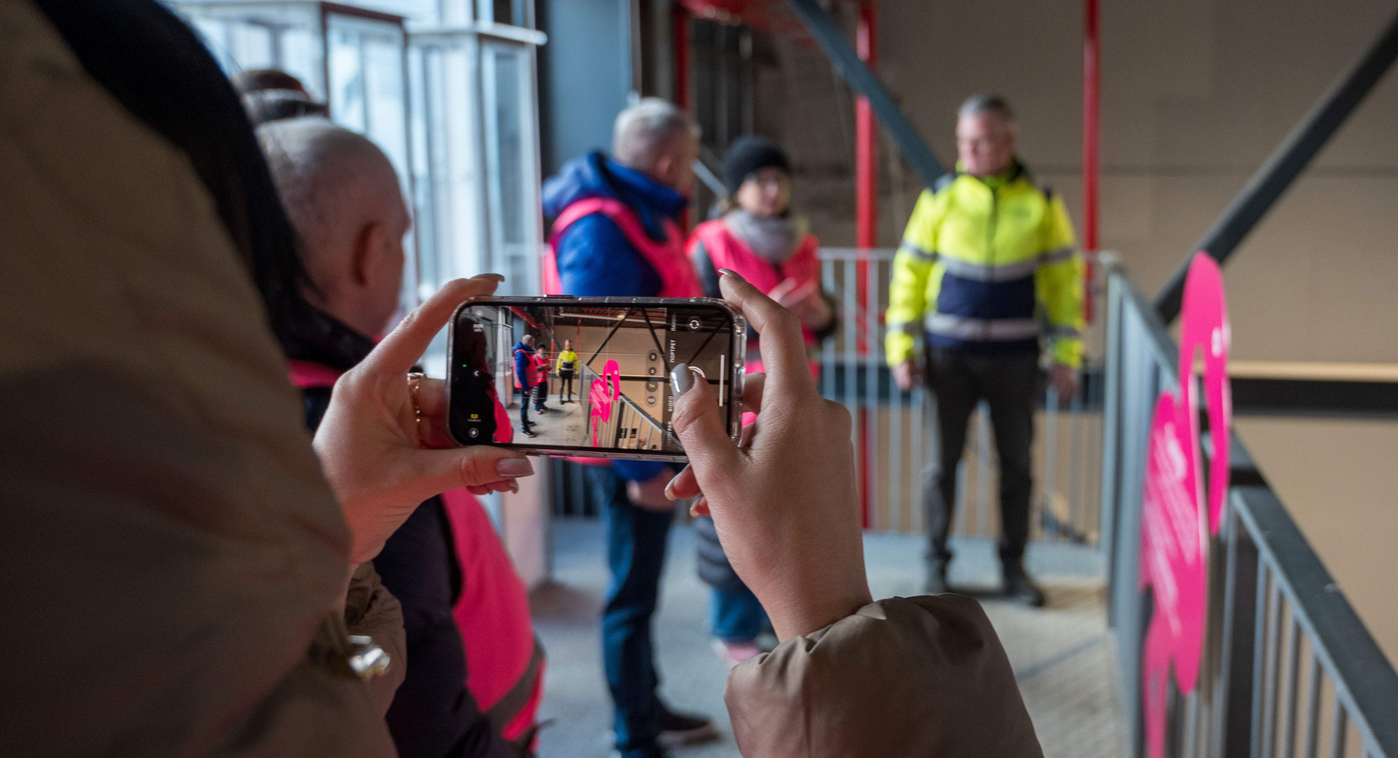 En person er i gang med at fotografere med en smartphone. På skærmen og i baggrunden ses en af platformene på Læringsbroen og flere personer i ført pink sikkerhedsveste står og taler sammen med en person i gul sikkerhedsjakke.