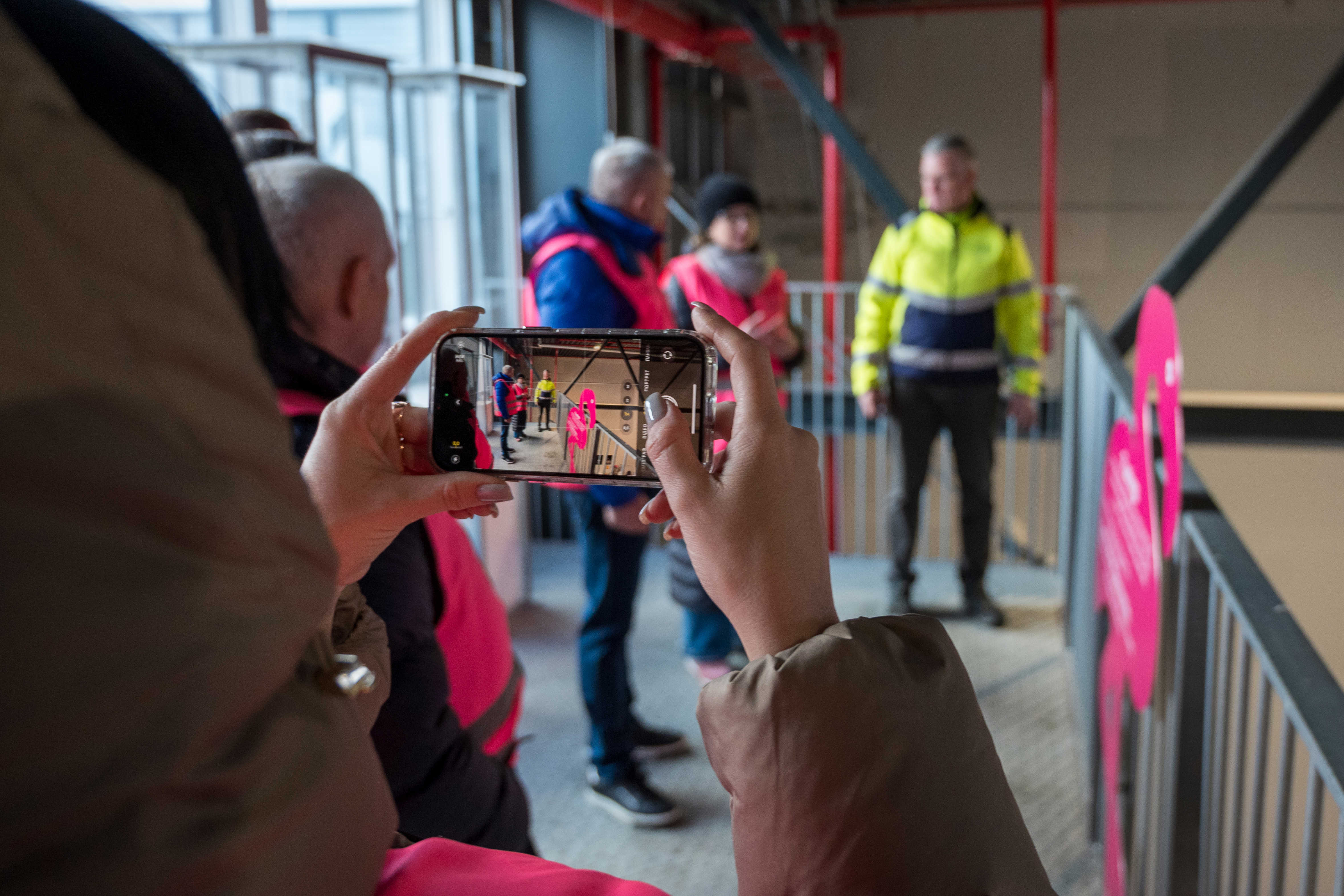 En person er i gang med at fotografere med en smartphone. På skærmen og i baggrunden ses en af platformene på Læringsbroen og flere personer i ført pink sikkerhedsveste står og taler sammen med en person i gul sikkerhedsjakke.
