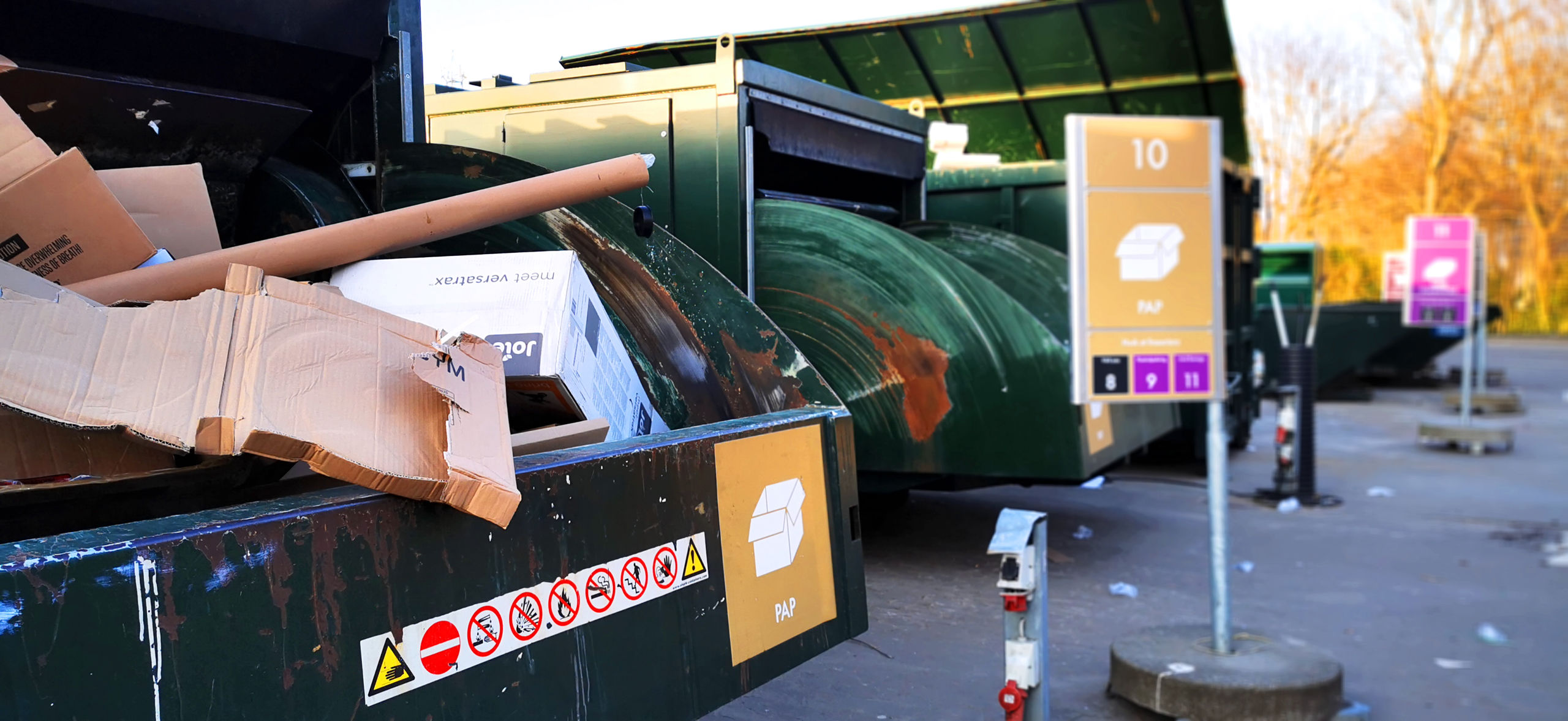 Genbrugsstation med store grønne containere til papaffald. En container i forgrunden er fyldt med papkasser og rør, og et skilt markerer området for papsortering. Flere containere til forskellige affaldsfraktioner ses i baggrunden.