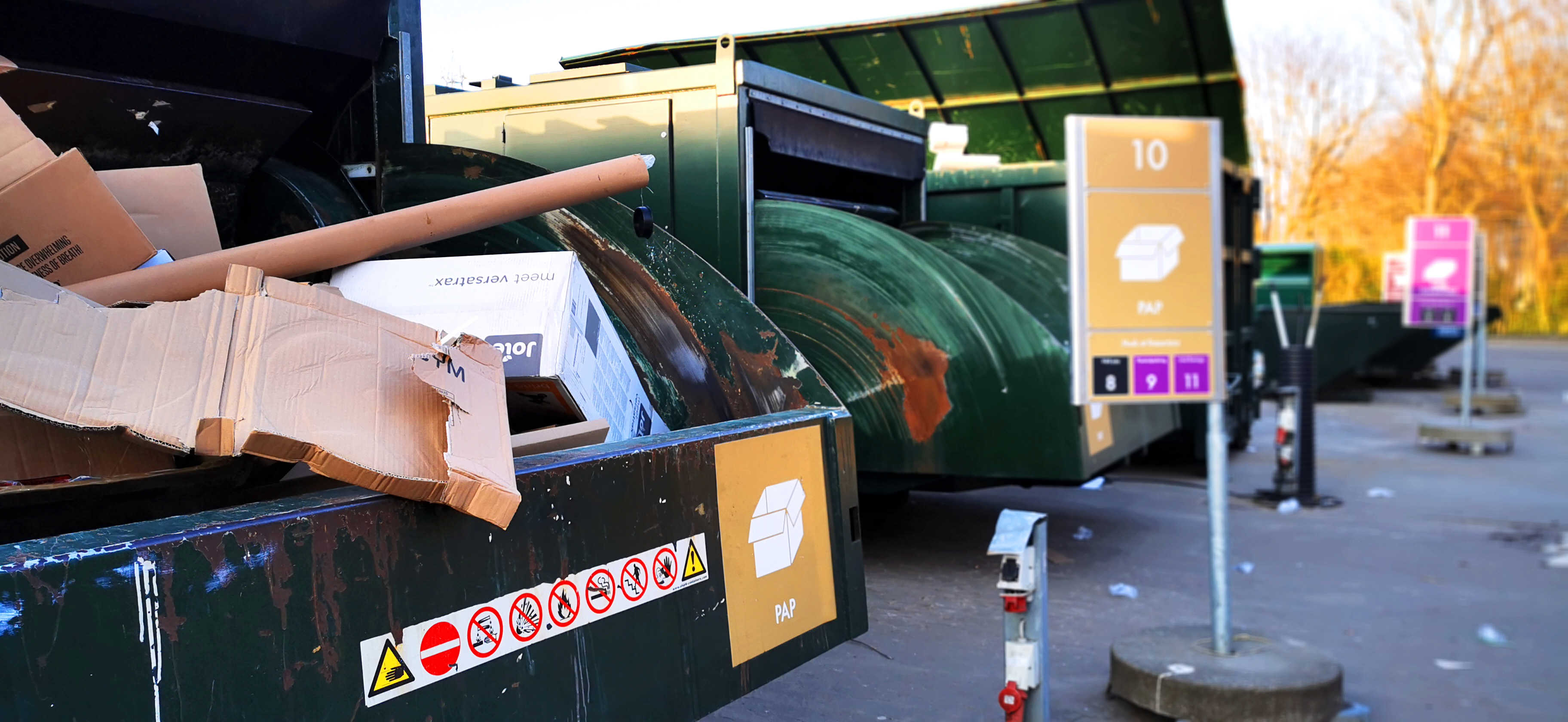 Genbrugsstation med store grønne containere til papaffald. En container i forgrunden er fyldt med papkasser og rør, og et skilt markerer området for papsortering. Flere containere til forskellige affaldsfraktioner ses i baggrunden.