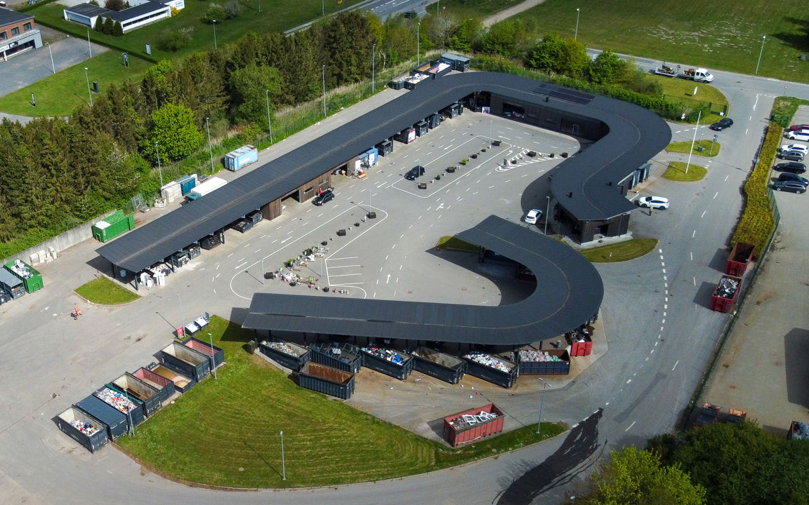 Oversigtsfoto af hele Give Genbrugsplads omgivet af grønne områder og vejnet. Billedet viser hele den buede bygning, kørselsretninger, containere og parkeringsområde.