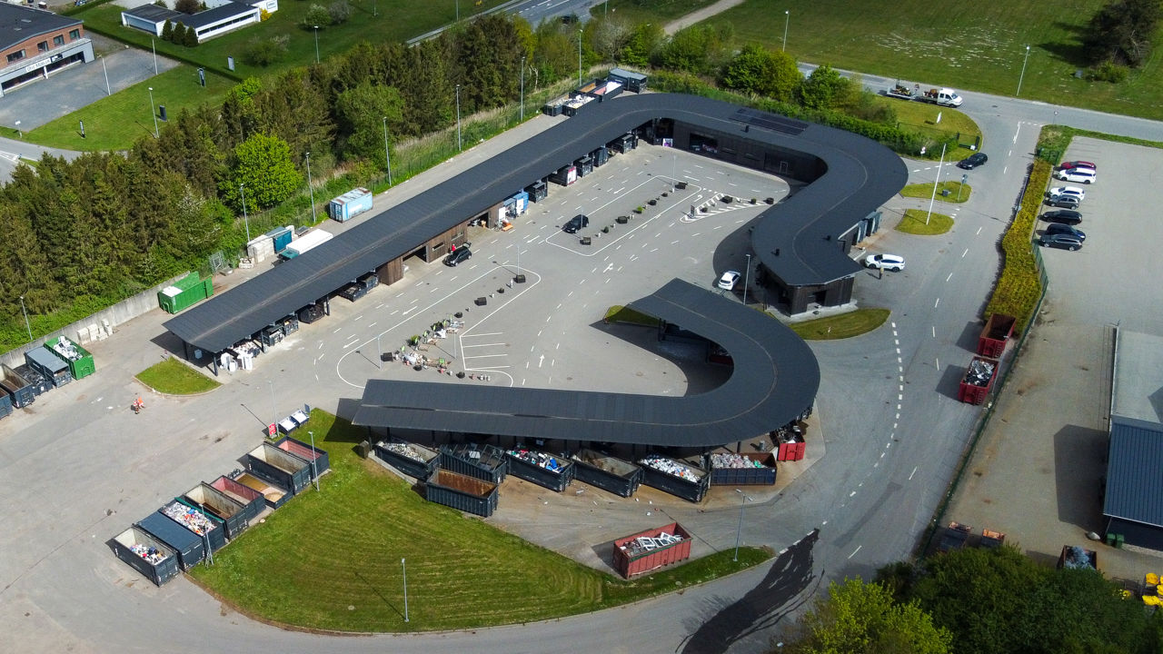 Oversigtsfoto af hele Give Genbrugsplads omgivet af grønne områder og vejnet. Billedet viser hele den buede bygning, kørselsretninger, containere og parkeringsområde.