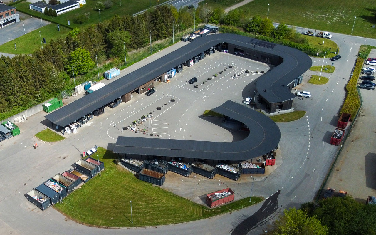 Oversigtsfoto af hele Give Genbrugsplads omgivet af grønne områder og vejnet. Billedet viser hele den buede bygning, kørselsretninger, containere og parkeringsområde.