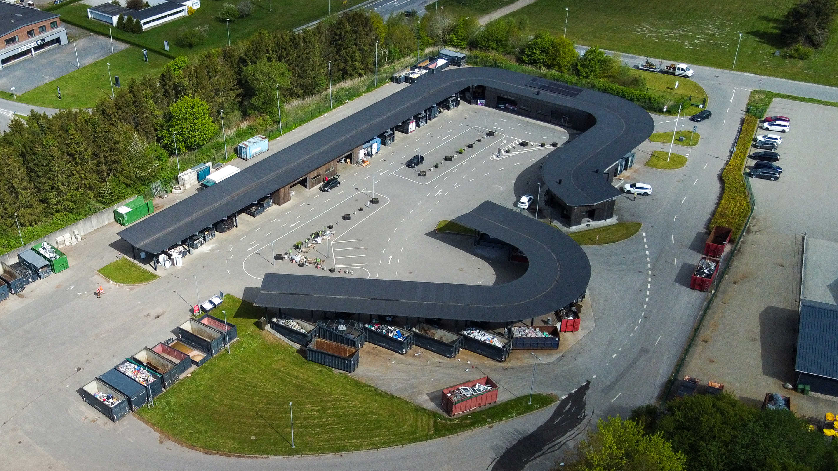 Oversigtsfoto af hele Give Genbrugsplads omgivet af grønne områder og vejnet. Billedet viser hele den buede bygning, kørselsretninger, containere og parkeringsområde.