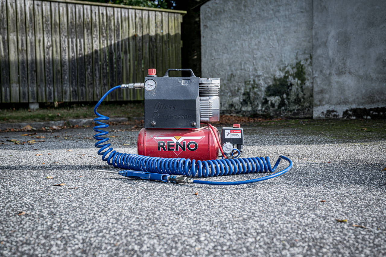 Kompressor placeret på asfalt foran et plankeværk.