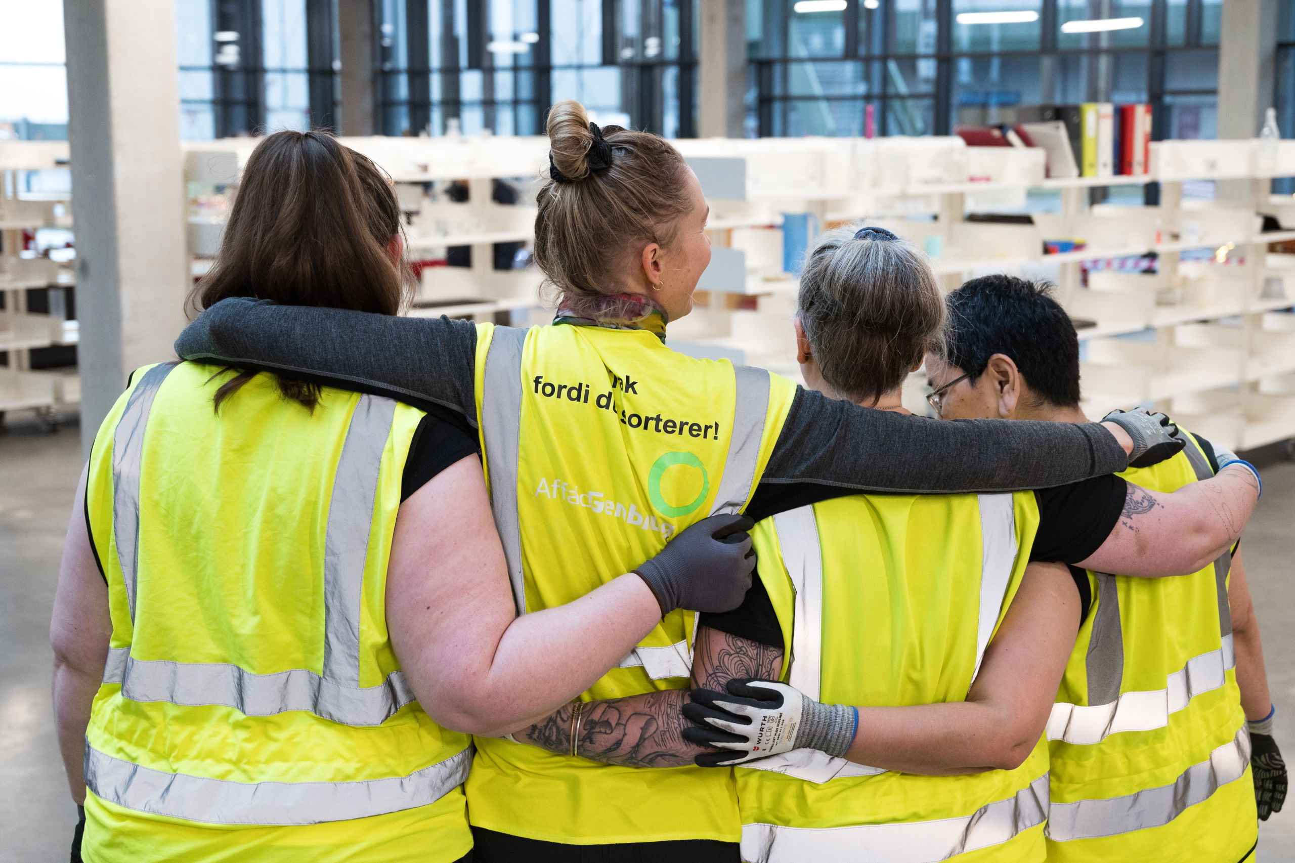 Fire medarbejdere iført gule refleksveste står sammen med armene om hinanden i Butikken på RessourceCenter Vejle (RCV). De arbejder med genbrug og genanvendelse som en del af et beskæftigelsesprojekt i samarbejde med Vejle Kommunes Jobcenter. Bag dem ses hylder med genbrugsvarer i butikslokalet.