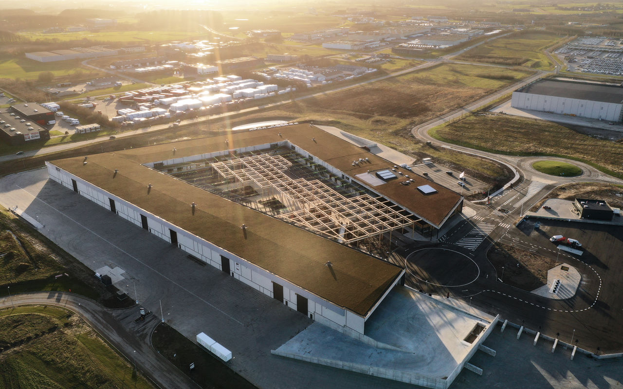 Dronefoto af RessourceCenter Vejle (RCV) med et grønt tag og solpaneler, omgivet af parkeringsplads og grønne områder, der viser centerets bæredygtige arkitektur og indre gårdstruktur.
