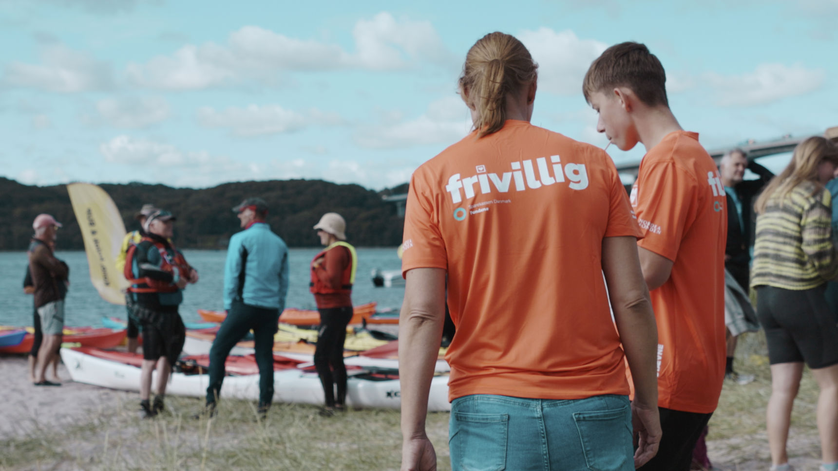 Frivillige i orange trøjer står ved Vejle Fjord med kajakker, mens en gruppe mennesker står og snakker