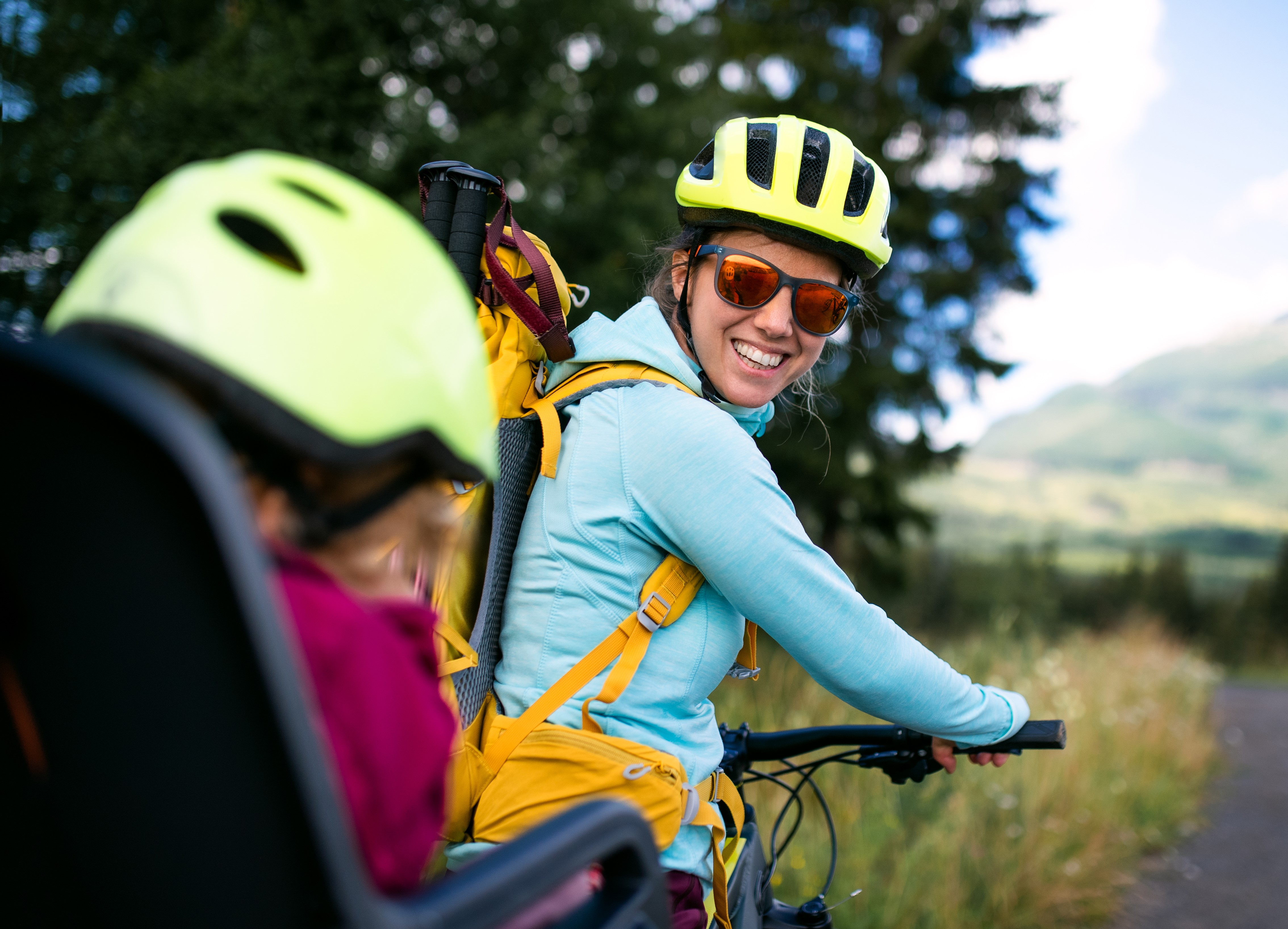 En smilende kvinde iført cykelhjelm og solbriller cykler med et barn i barnesæde bagpå. De er omgivet af grøn natur