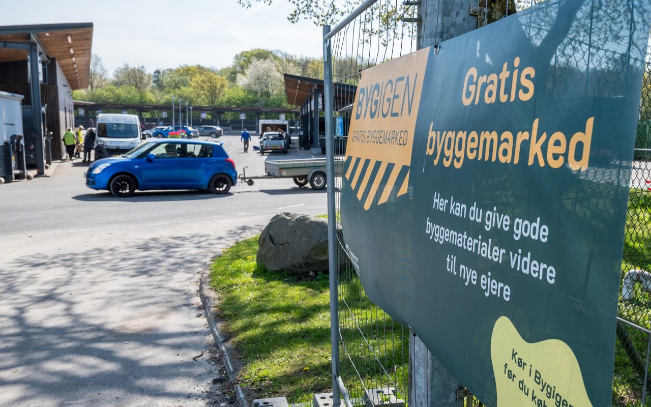 Skilt ved indgangen til BygIgen ved Børkop Genbrugsplads. Skiltet annoncerer "Gratis byggemarked" og inviterer borgere til at aflevere eller afhente gode byggematerialer, der kan genbruges.