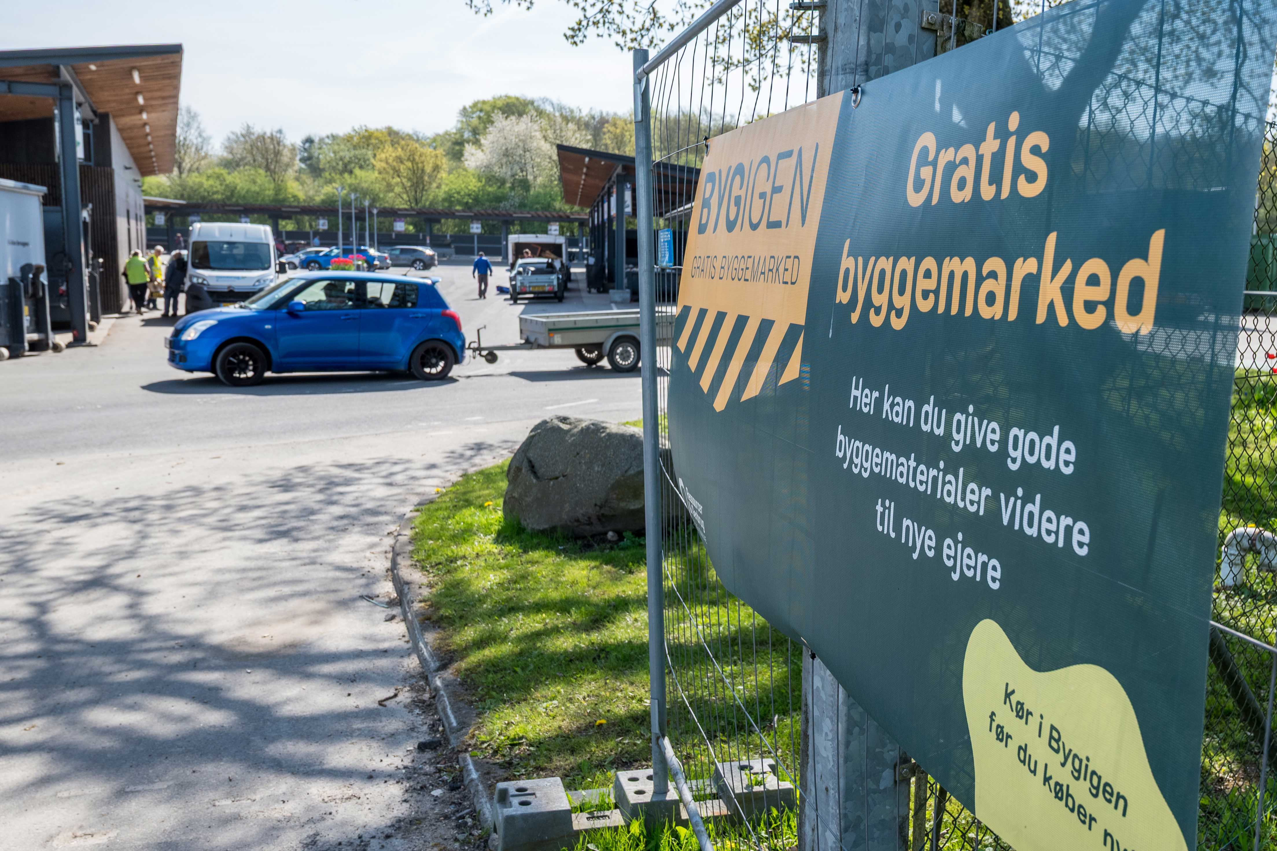 Skilt ved indgangen til BygIgen ved Børkop Genbrugsplads. Skiltet annoncerer "Gratis byggemarked" og inviterer borgere til at aflevere eller afhente gode byggematerialer, der kan genbruges.