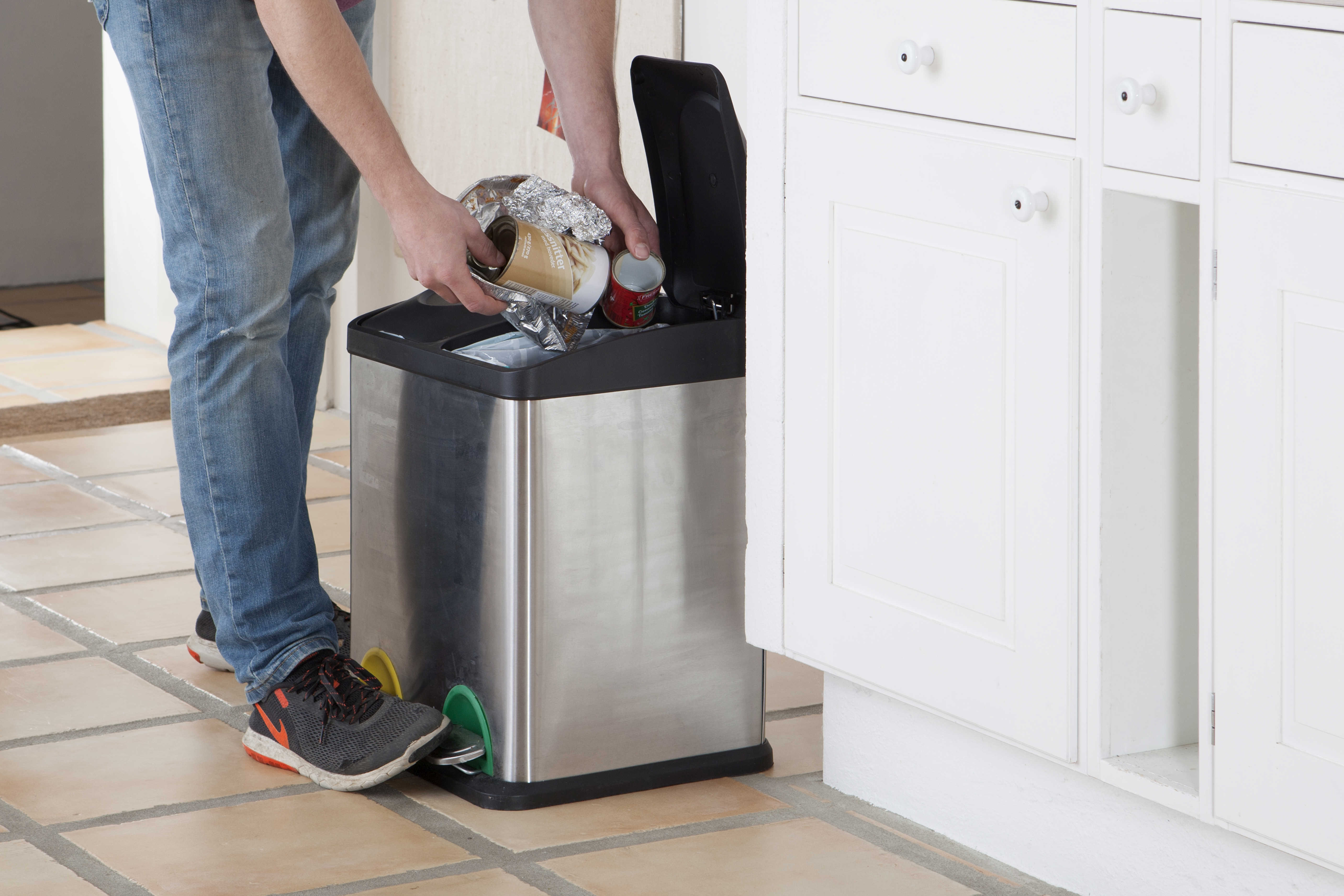 Person i jeans og sneakers smider affald i en skraldespand af rustfrit stål i et køkken med hvide skabe og flisegulv.