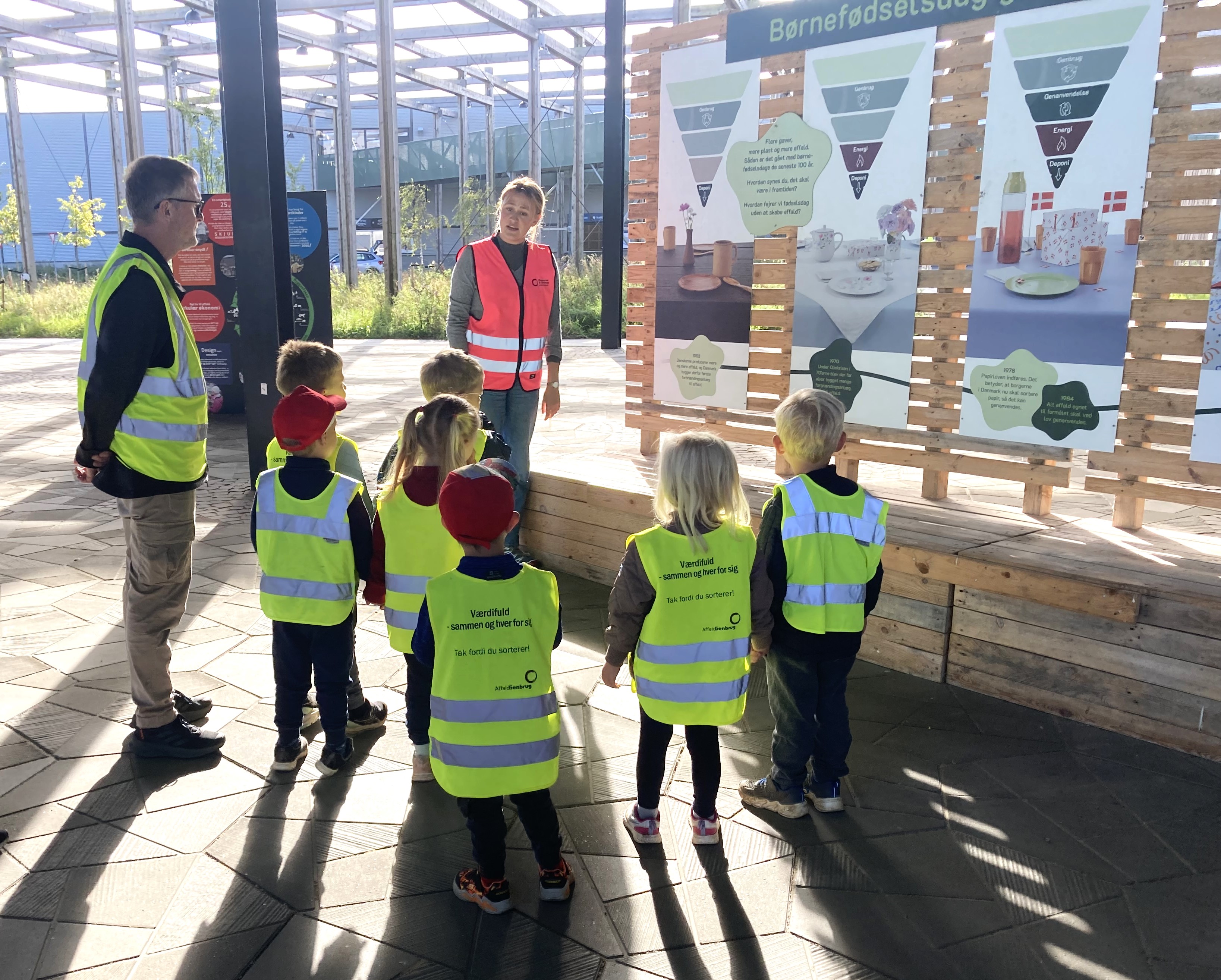Genbrugsformidler iført pink sikkerhedsvest på Torvet ved RCV sammen med en gruppe børnehavebørn, der bliver vist rundt.