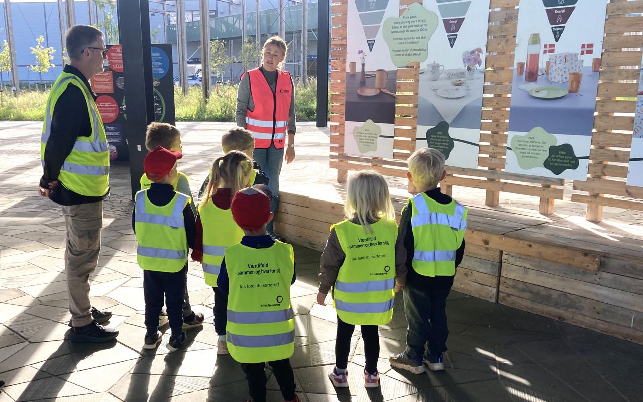 Genbrugsformidler iført pink sikkerhedsvest på Torvet ved RCV sammen med en gruppe børnehavebørn, der bliver vist rundt.