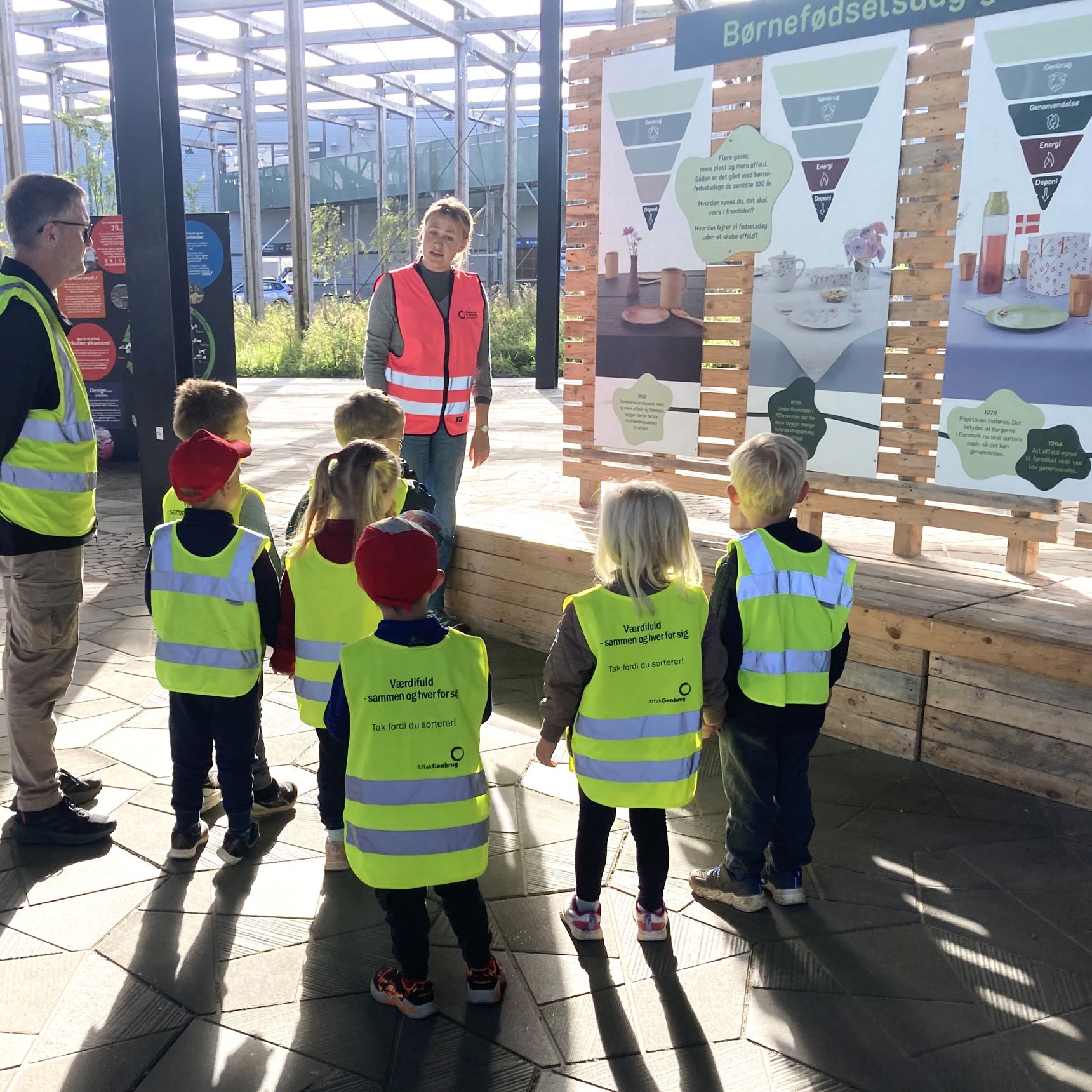 Genbrugsformidler iført pink sikkerhedsvest på Torvet ved RCV sammen med en gruppe børnehavebørn, der bliver vist rundt.