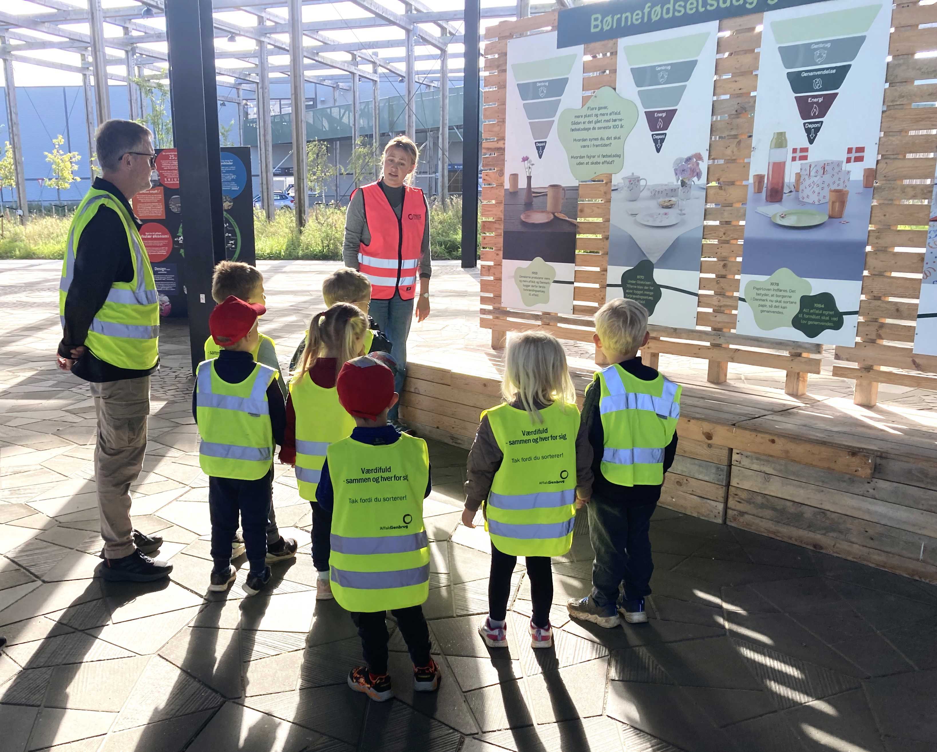 Genbrugsformidler iført pink sikkerhedsvest på Torvet ved RCV sammen med en gruppe børnehavebørn, der bliver vist rundt.