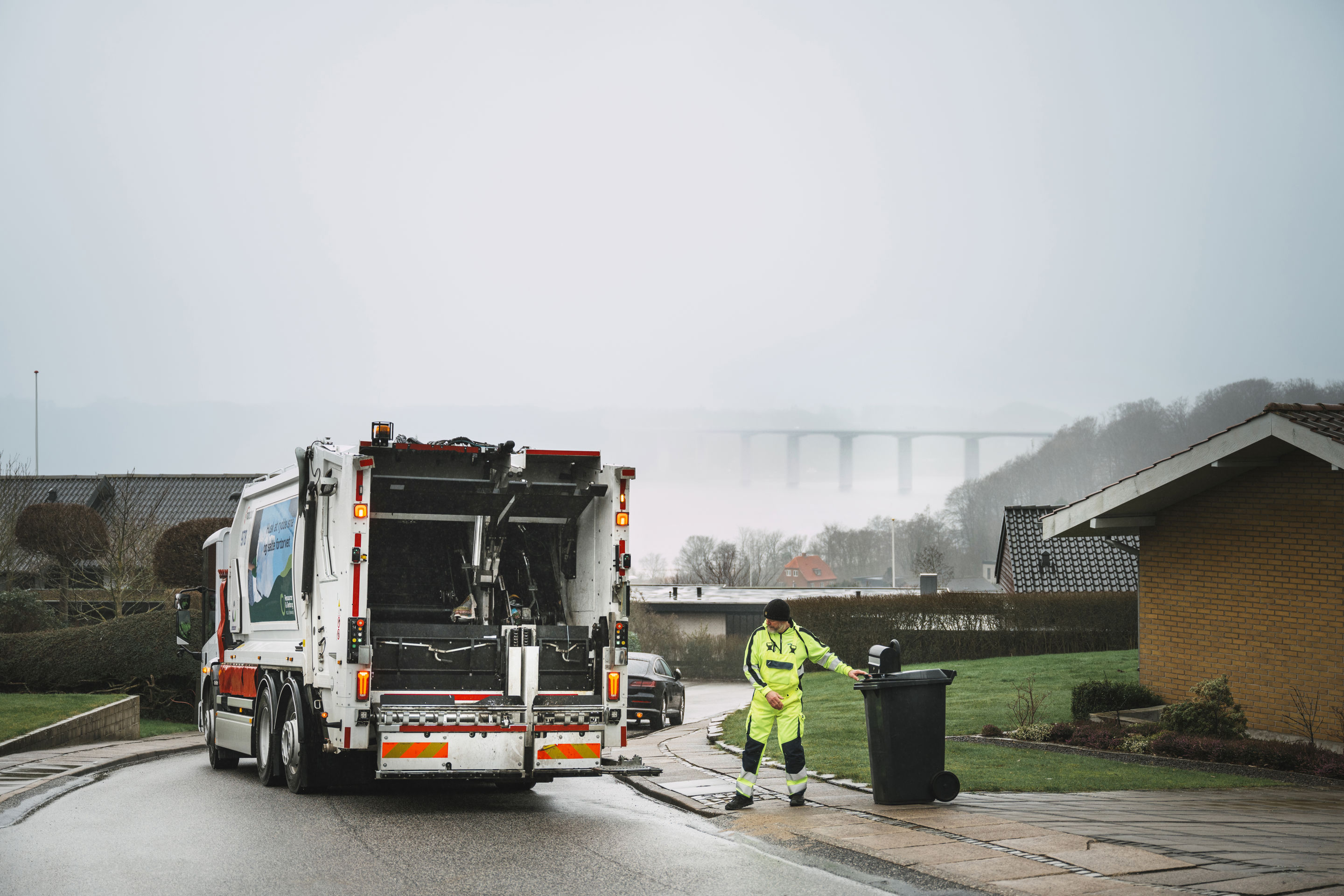 Bagenden af en skraldebil under arbejdet, med udsigt over et boligområde.