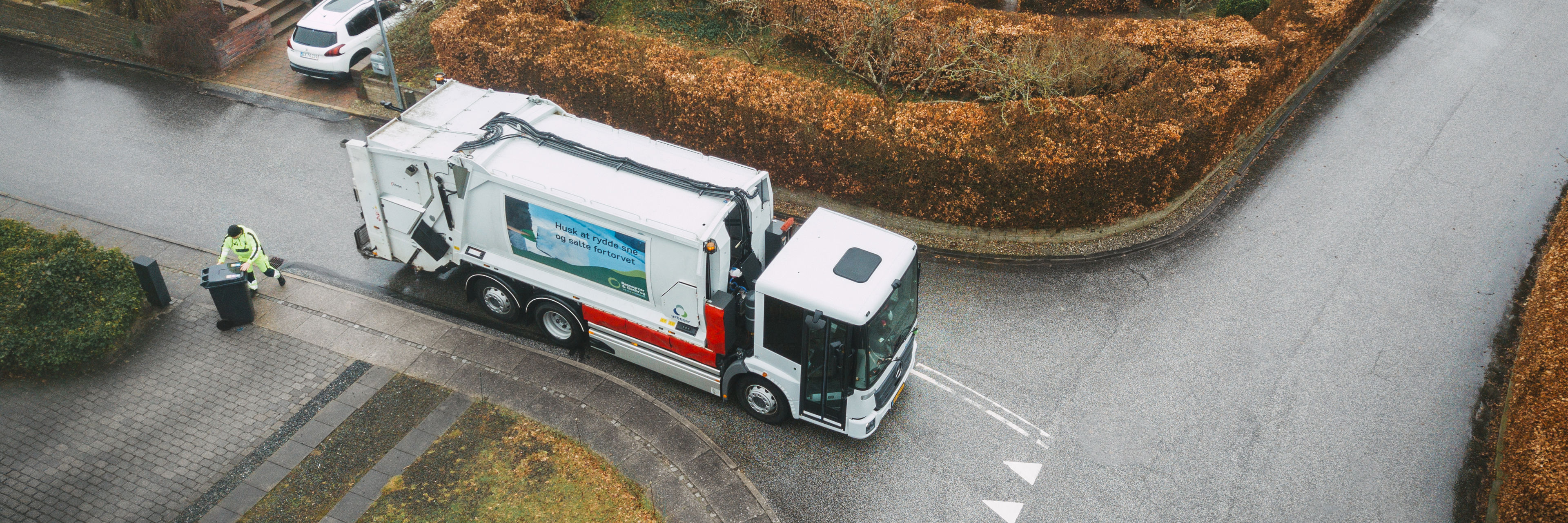 En skraldebil, der kører rundt i et boligområde med velplejede hække, mens en skraldemand tømmer en affaldsbeholder