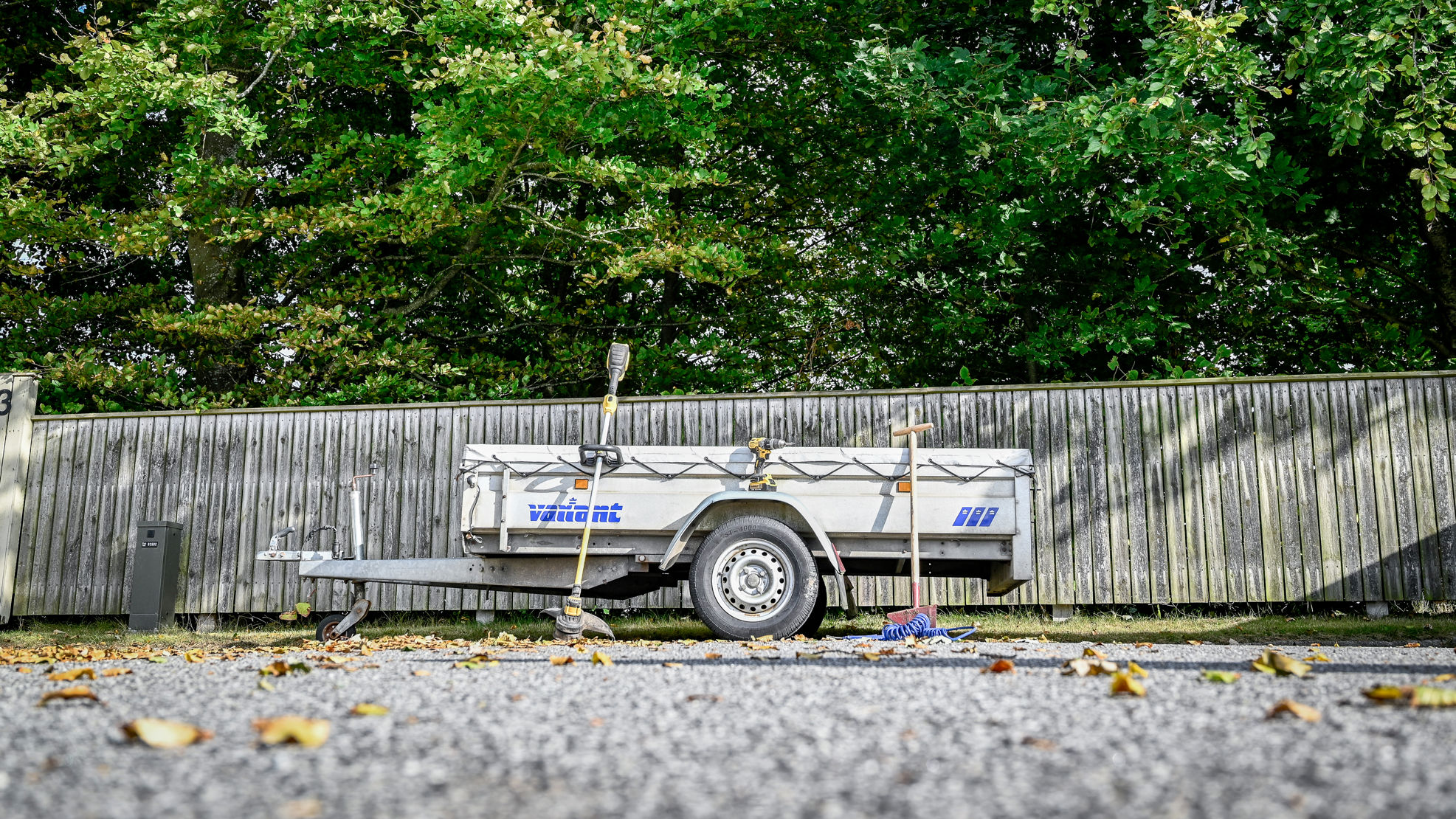 Anhænger parkeret på villavej med forskellige haveredskaber og værktøj omkring. Bagved ses stakit og grønne trækroner.