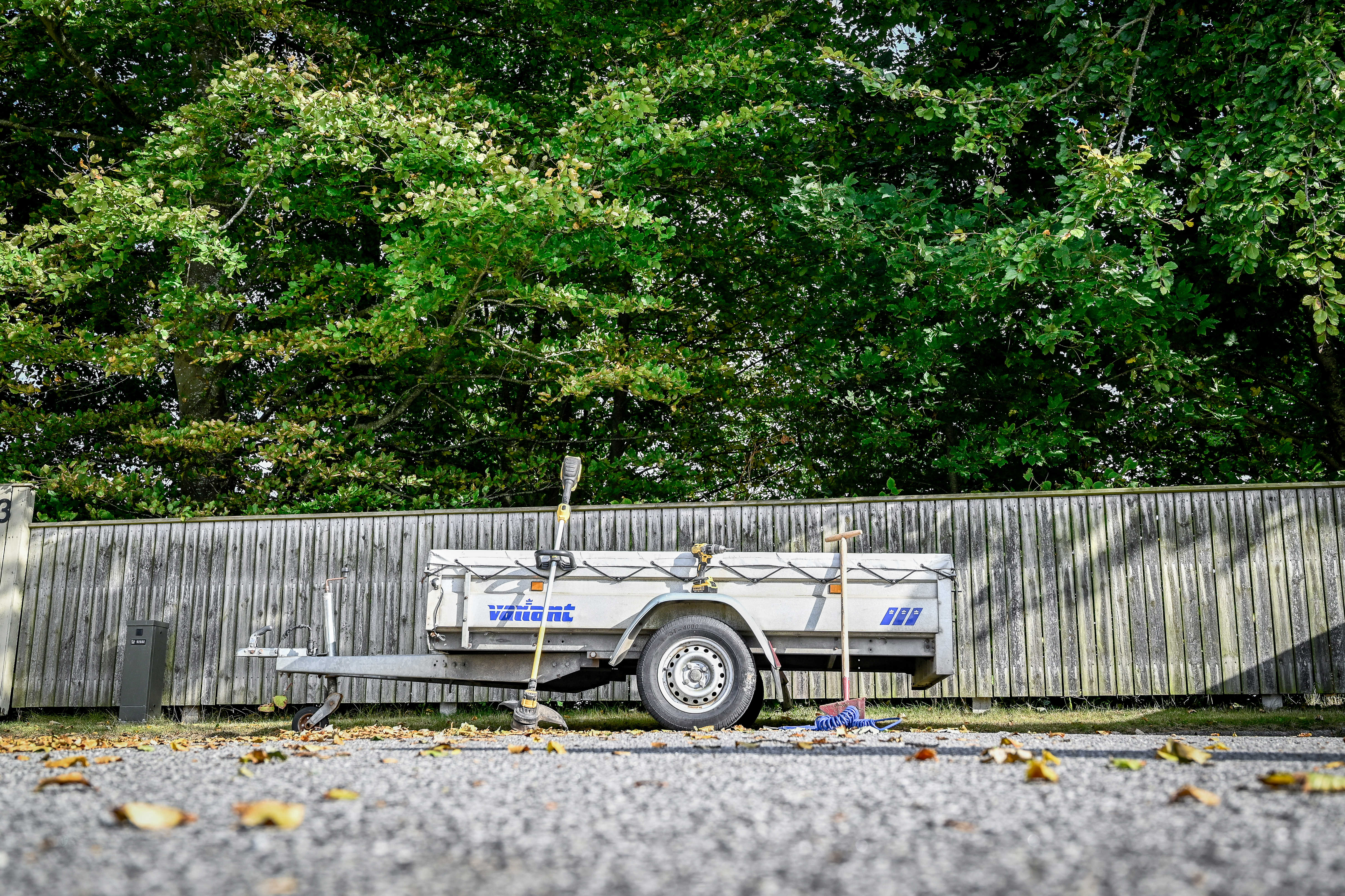 Anhænger parkeret på villavej med forskellige haveredskaber og værktøj omkring. Bagved ses stakit og grønne trækroner.